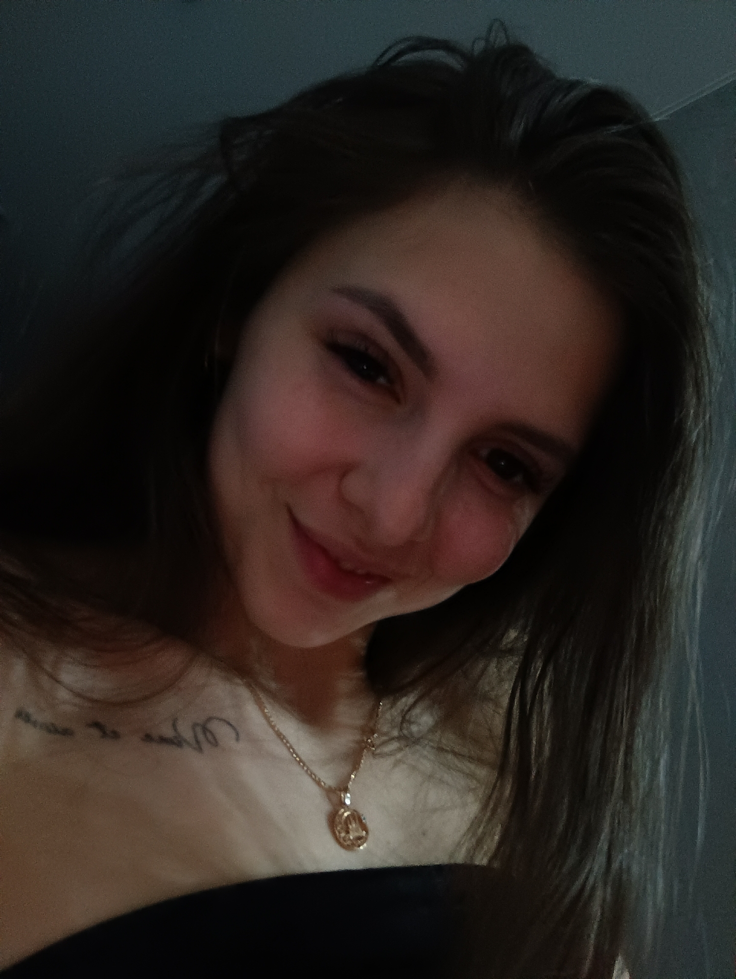 A woman with long, dark hair smiles at the camera. She wears a gold necklace with a round pendant and has a tattoo on her upper chest. The background appears to be a dimly lit interior. The image does not provide information about the location in the city of Chaikovsky, Russia.