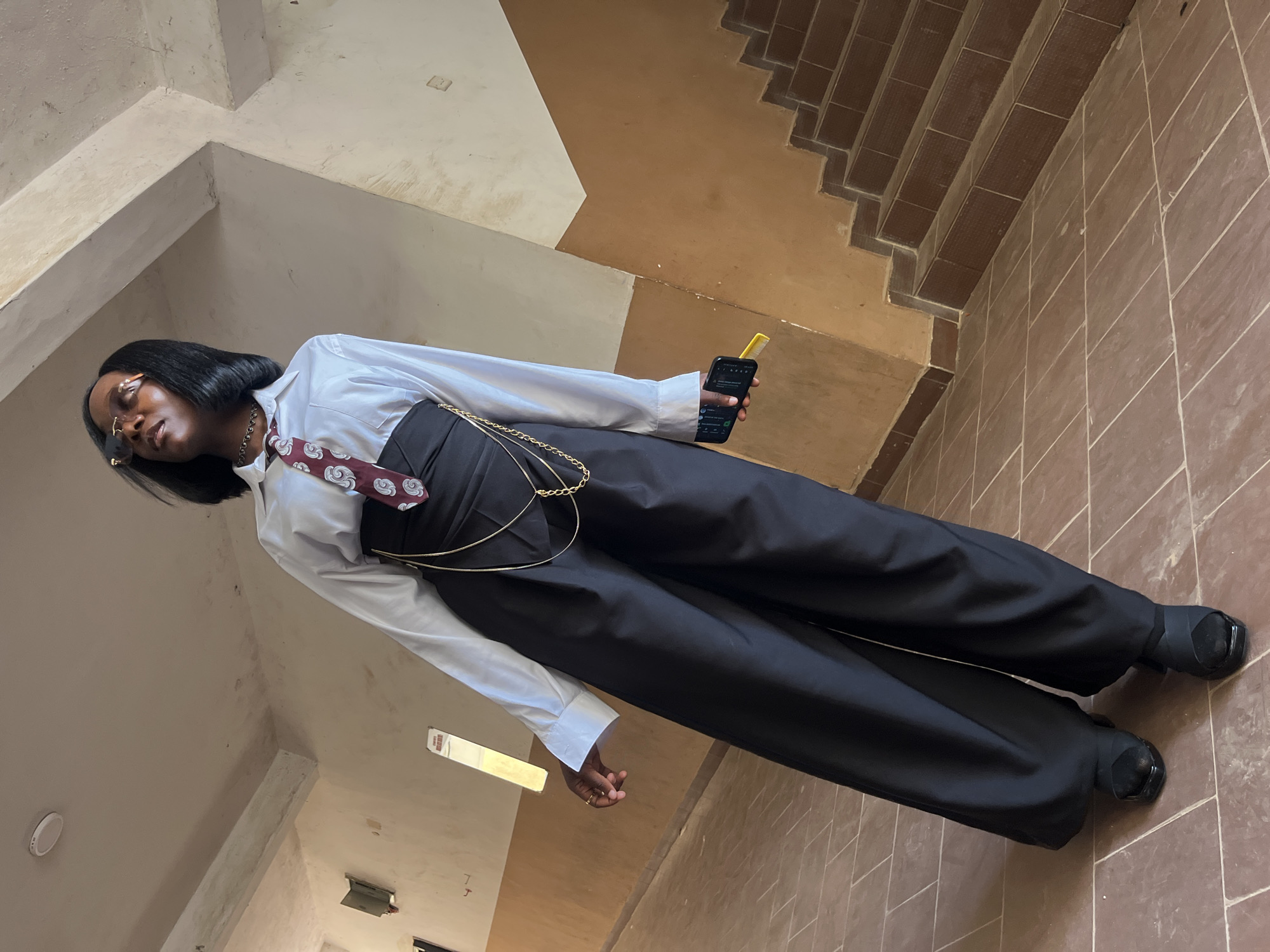 A person stands in a hallway with a tiled floor and stairwell to the right. The individual is wearing a white collared shirt, a dark tie with a circular pattern, a black corset-like garment, and wide-legged black pants. They are also wearing black shoes and sunglasses, and are holding a black smartphone in their right hand. The hallway appears to be an interior space with painted walls and a concrete ceiling. The lighting suggests daytime. The location is identified as the city of Jos, Nigeria.