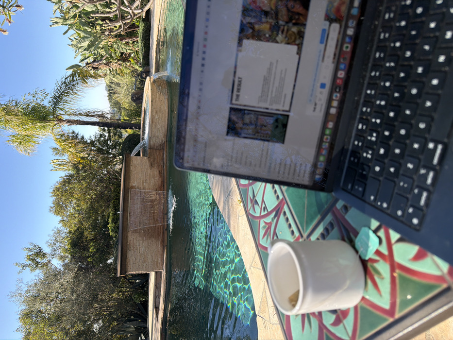 A laptop sits on a patterned table next to a pool with a waterfall feature. Palm trees and other greenery are visible in the background against a clear blue sky. A white cup with a small object inside is on the table next to the laptop, along with a blue stone. The laptop screen displays a document titled "THE RESULT." The scene suggests an outdoor workspace by the pool.