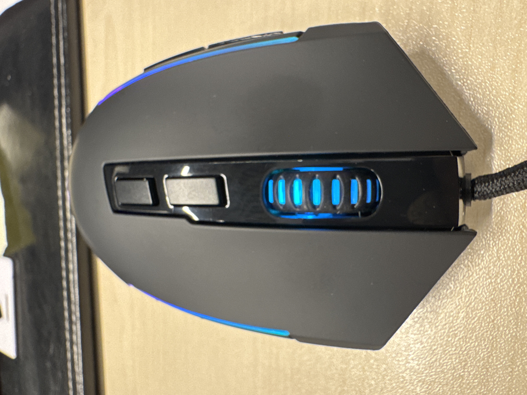 An overhead, close-up shot shows a black computer mouse with blue LED lighting. The mouse is situated on a tan, textured surface, likely a desk. The lighting on the mouse is most prominent around the scroll wheel, which is also blue, and along the outer edges of the mouse's casing. The mouse's design is angular, with two primary buttons at the top and a textured scroll wheel in the center. A black braided cable extends from the front of the mouse. The background consists of a blurred, dark surface that appears to be part of a computer setup. The scene is indoors and appears to be a typical office or home workspace. There are no people or animals visible. The city of Almaty, Kazakhstan, is not visually represented in this image.
