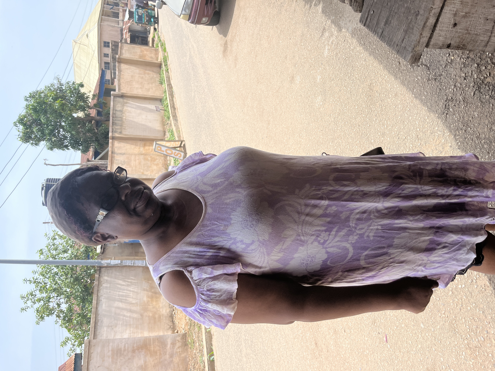 A woman is seen standing on a paved road in Jos, Nigeria. She wears a light purple floral dress and sunglasses. Behind her are buildings and trees, and a partially visible vehicle is in the background. The lighting suggests it is daytime.