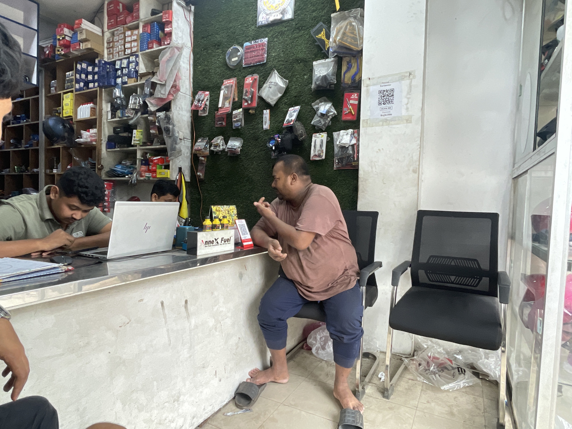The photo shows the interior of a small shop, potentially an auto parts or motorcycle parts store, in Dhaka, Bangladesh. A man sits on a chair behind a counter, wearing a brown t-shirt and blue pants. He is gesturing with his hands as if in conversation. Another man, wearing a green shirt, is sitting at the counter, looking at a laptop computer. Various shelves filled with merchandise line the walls. The walls are covered in green decorative artificial foliage on which a variety of motorcycle parts for sale are hanging. There is a QR code placed on the wall, possibly related to the shop. The shop appears to be small and cluttered.