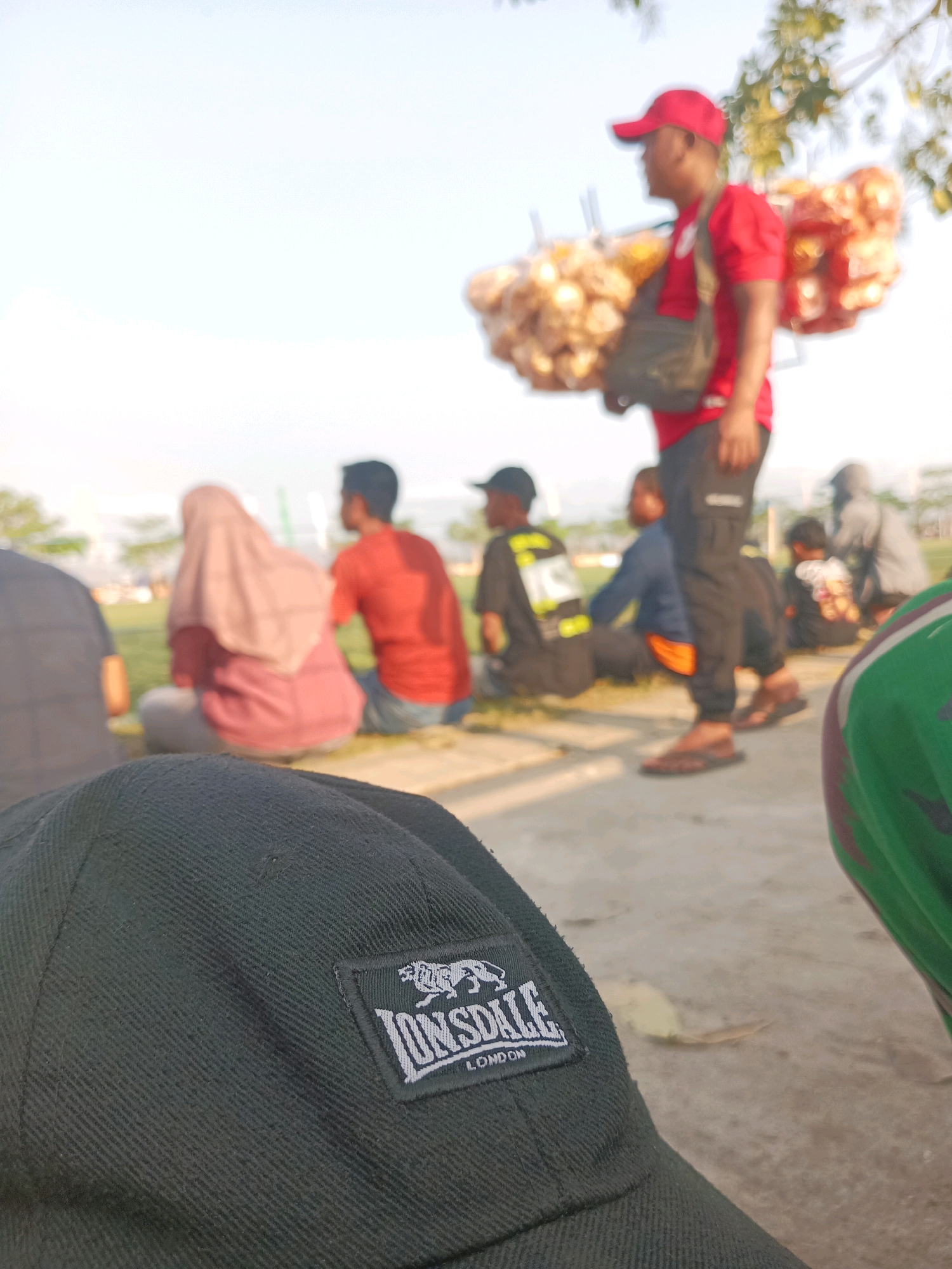 A close-up of a black cap with a white Lonsdale logo is in the foreground, obscuring the lower portion of the image. In the background, a vendor wearing a red cap and shirt carries snacks on a rack over his shoulder, walking on a paved path. Several people are seated on the ground in the middle ground, including a person wearing a pink headscarf and a person in a red shirt. The scene is outdoors, with trees and foliage visible in the distance under a bright sky. The image appears to be taken in the city of Tawing, Indonesia.