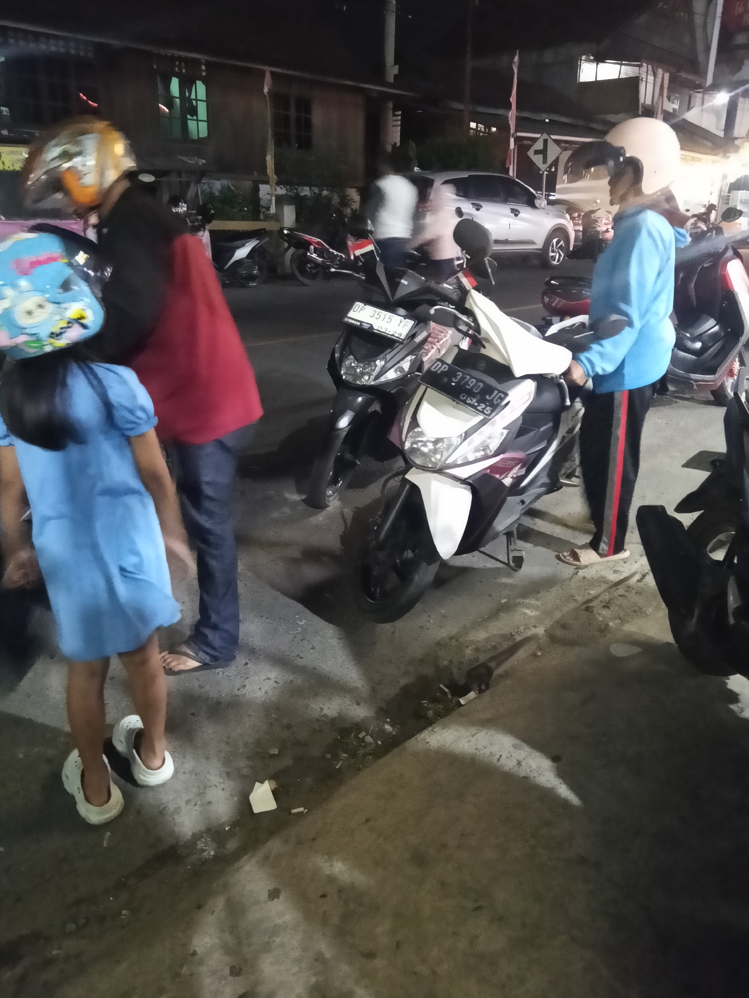 The image depicts a street scene at night in Makale, Indonesia. In the foreground, a young girl wearing a blue dress and white shoes stands next to an adult wearing dark pants and a red shirt. The girl is facing away from the camera, and the adult is partially obscured. Several motorcycles are parked on the street, one of which is white and maroon with a license plate reading "DP 3790 JG." Another motorcycle, black and silver, is parked behind it. In the background, a silver car is visible, as well as buildings with illuminated windows and streetlights. The overall impression is a busy, urban environment at night.