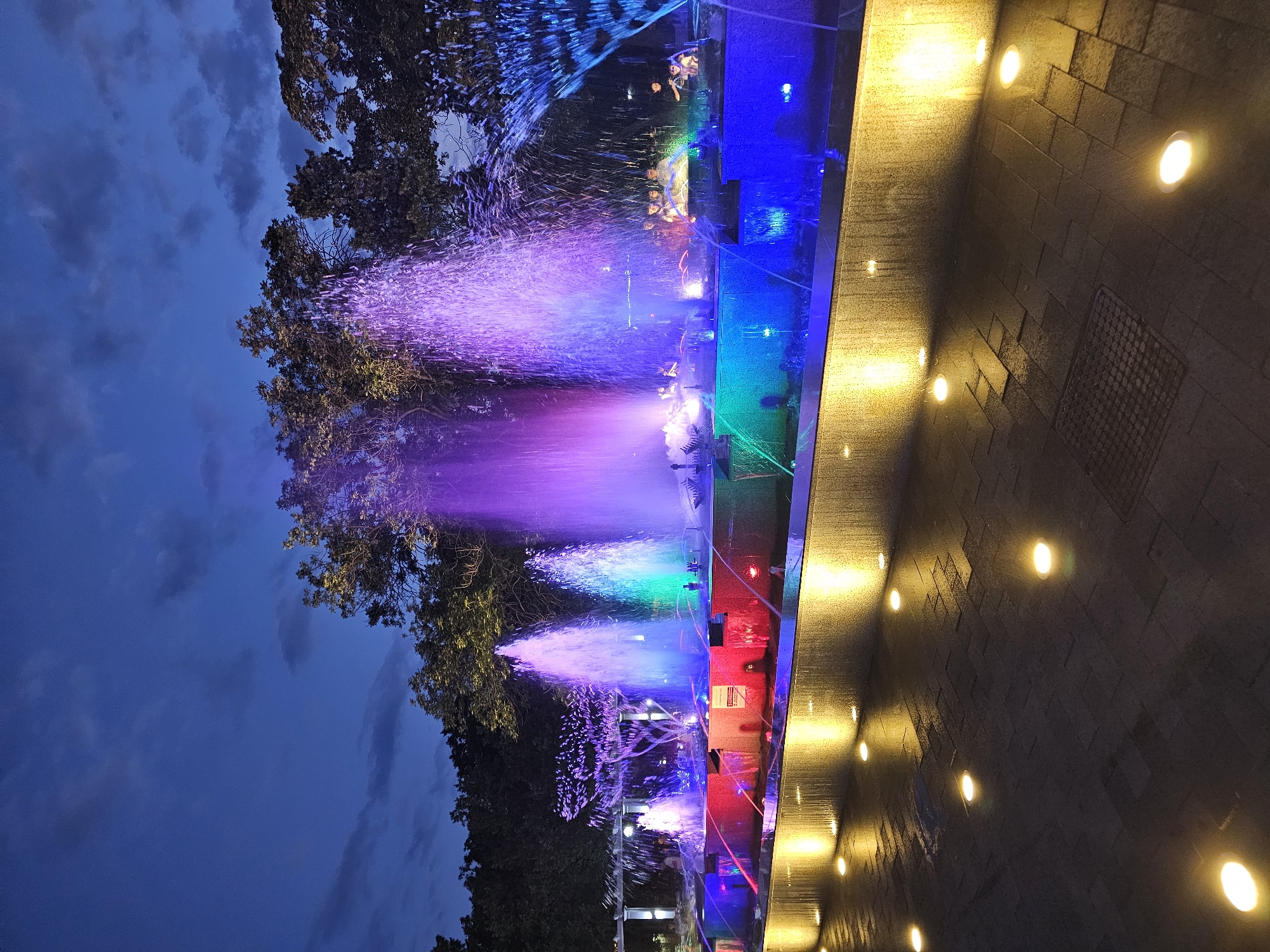 The image shows a large, multi-tiered water fountain illuminated with vibrant colored lights at dusk. The fountain is the central focus, with water jets creating various patterns and reaching different heights. The water is lit in shades of purple, pink, green, and blue, creating a dynamic visual effect.
In the background, silhouetted trees form a dark canopy against the twilight sky. The sky itself is a deep blue, with some faint clouds visible.
In the foreground, the paved ground is illuminated by a series of warm-toned, circular lights embedded in the surface, casting a soft glow and creating reflections on the wet pavement.
There are several people visible around the fountain, some standing and observing, while others appear to be seated or interacting with the water features. They are indistinct due to the low light conditions. The setting appears to be an urban park or public plaza.
The location is the Fountain of Youth in Cherkessk, Russia.