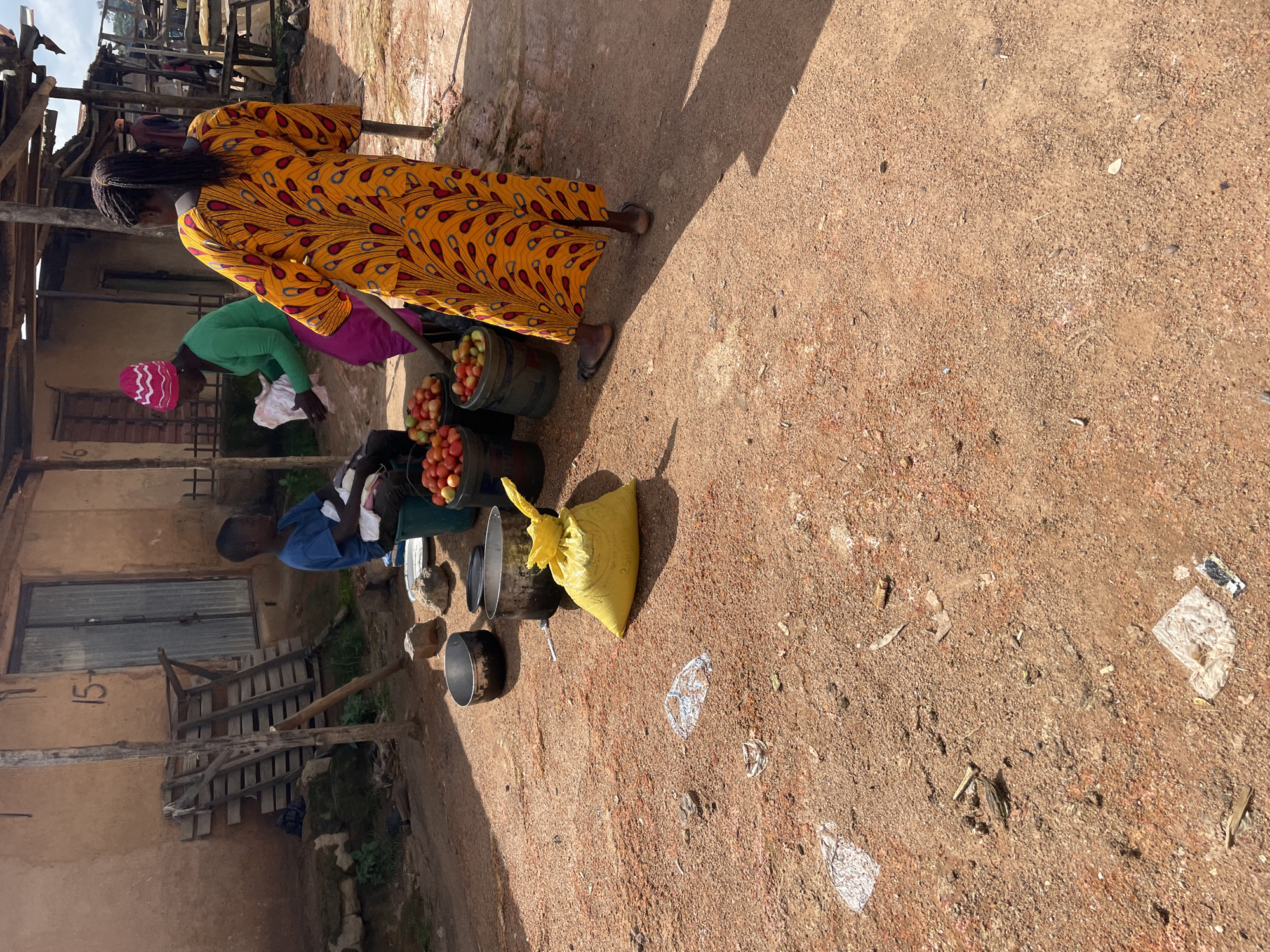 In Wang, Nigeria, a man sits on a green plastic bucket on a dirt ground outside a building. He is wearing a blue t-shirt and dark pants. A woman in a yellow dress with a vibrant pattern stands to his right, facing away from the camera. She is engaged in a transaction with another woman wearing a green long-sleeved shirt and a pink knitted hat. The second woman is holding a white cloth and appears to be offering it to the first woman. Several buckets filled with tomatoes are visible, along with large metal pots and a bright yellow bag. The ground is covered in reddish-brown dirt and scattered debris, including plastic and organic matter. The background features a rustic building with a corrugated metal door and wooden structures.