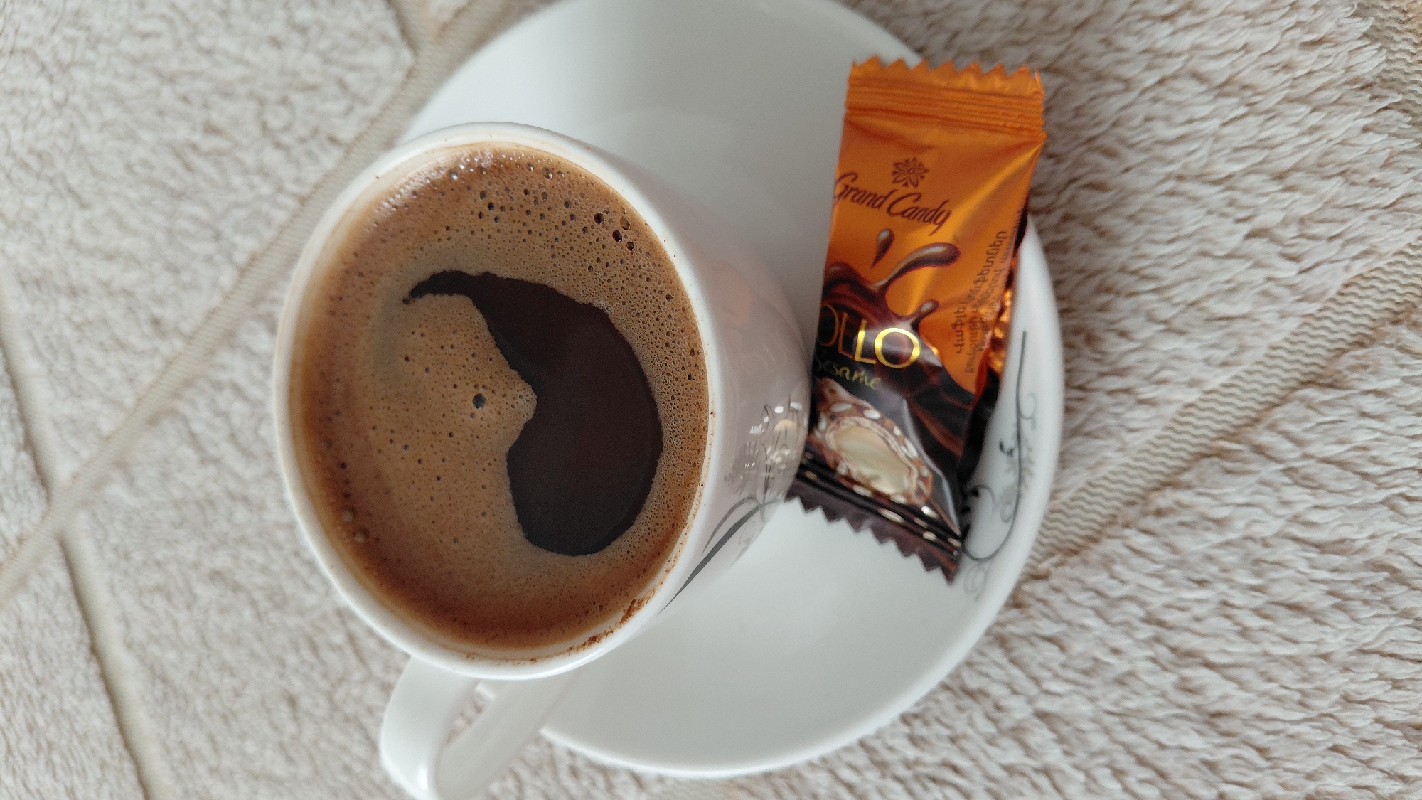 A close-up, top-down shot shows a white cup of coffee with a foam art design on a saucer. Next to the cup lies a chocolate candy wrapper. The scene is set on a textured, cream-colored surface, possibly a tablecloth. The location is Abovyan, Armenia.