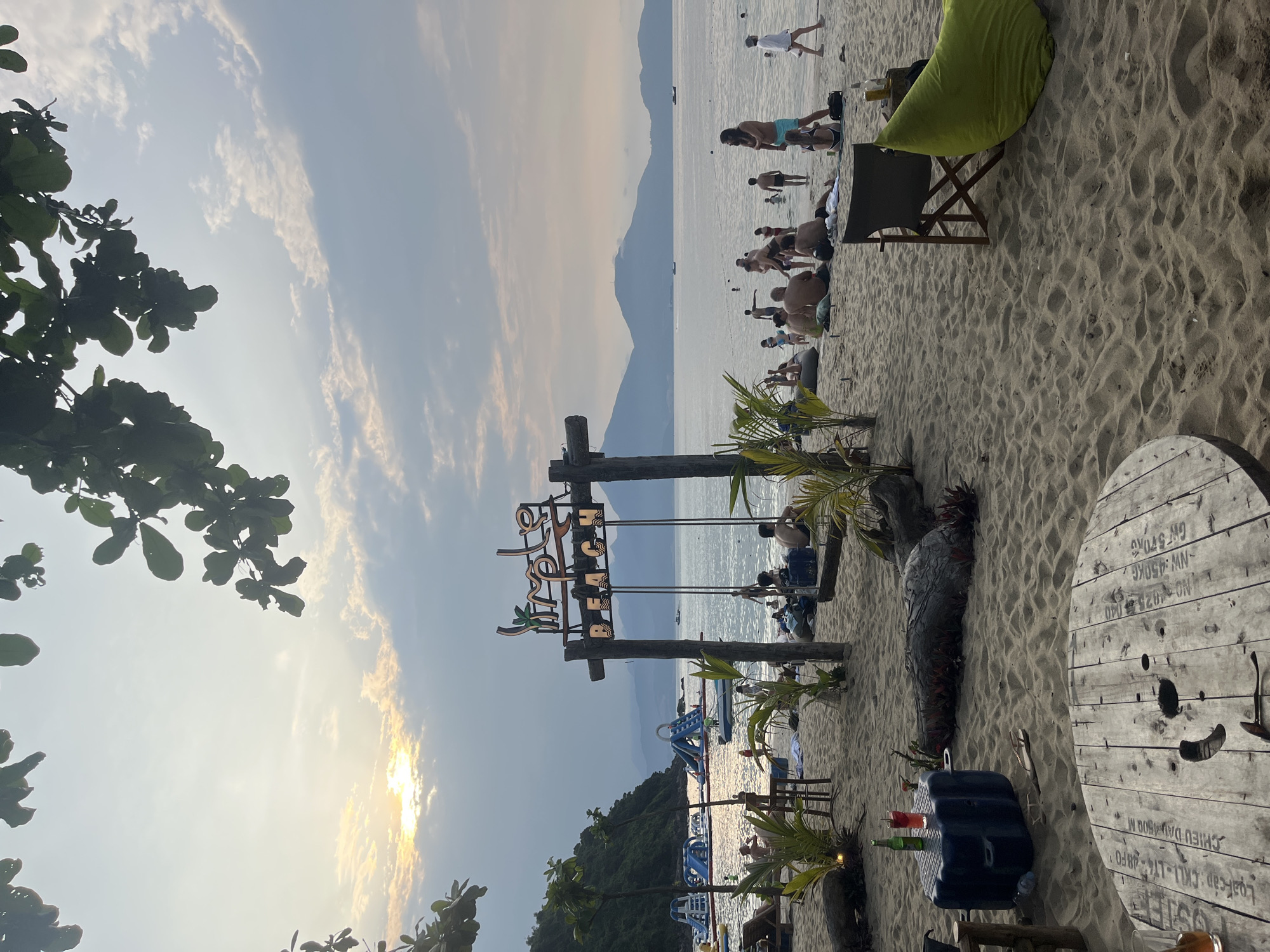 The image shows a beach scene in Đà Nẵng, Vietnam at sunset. The view is from an elevated position, looking down onto the sandy beach. The "Lazy Beach" sign is visible, indicating the location. Several people are on the beach; some are in the water, others are sunbathing or walking along the shore. Inflatable structures are present in the water near the shore. A large cable reel is used as a table, and there are chairs and a beanbag. A black trunk or cooler is next to the cable reel table with assorted drinks on it. Trees line part of the beach area. Mountains are visible in the background across the water.