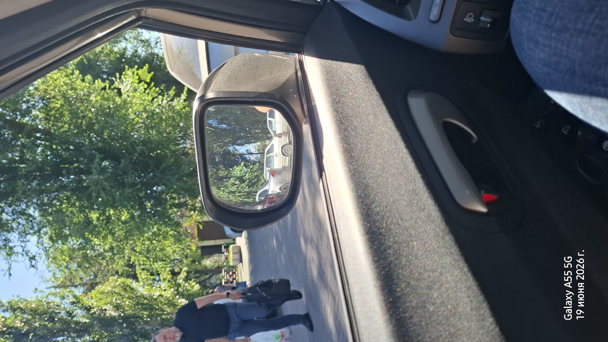 The image, taken from inside a car in Rostov-on-Don, Russia, shows a view through the open driver's side window. The side-view mirror reflects parked cars and green trees under a clear sky, suggesting it's daytime with fair weather. A person, wearing a dark shirt and blue jeans, is seen walking on a paved walkway in the reflection of the side-view mirror, carrying a black bag. The interior of the car is visible, including the door handle and part of the dashboard. The overall scene is one of everyday life, possibly a parking area near a park or residential zone. The metadata at the bottom right corner indicates the photo was taken on June 19, 2026, with a Galaxy A55 5G phone.