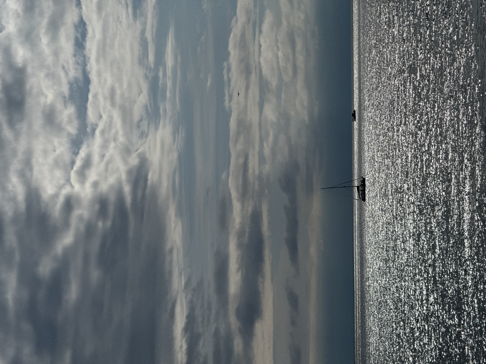 The image shows a body of water reflecting sunlight with a sailboat visible on the water. The sky is filled with clouds. The location is not discernible from the image content. No information about the city of Sirius, Russia, is evident.