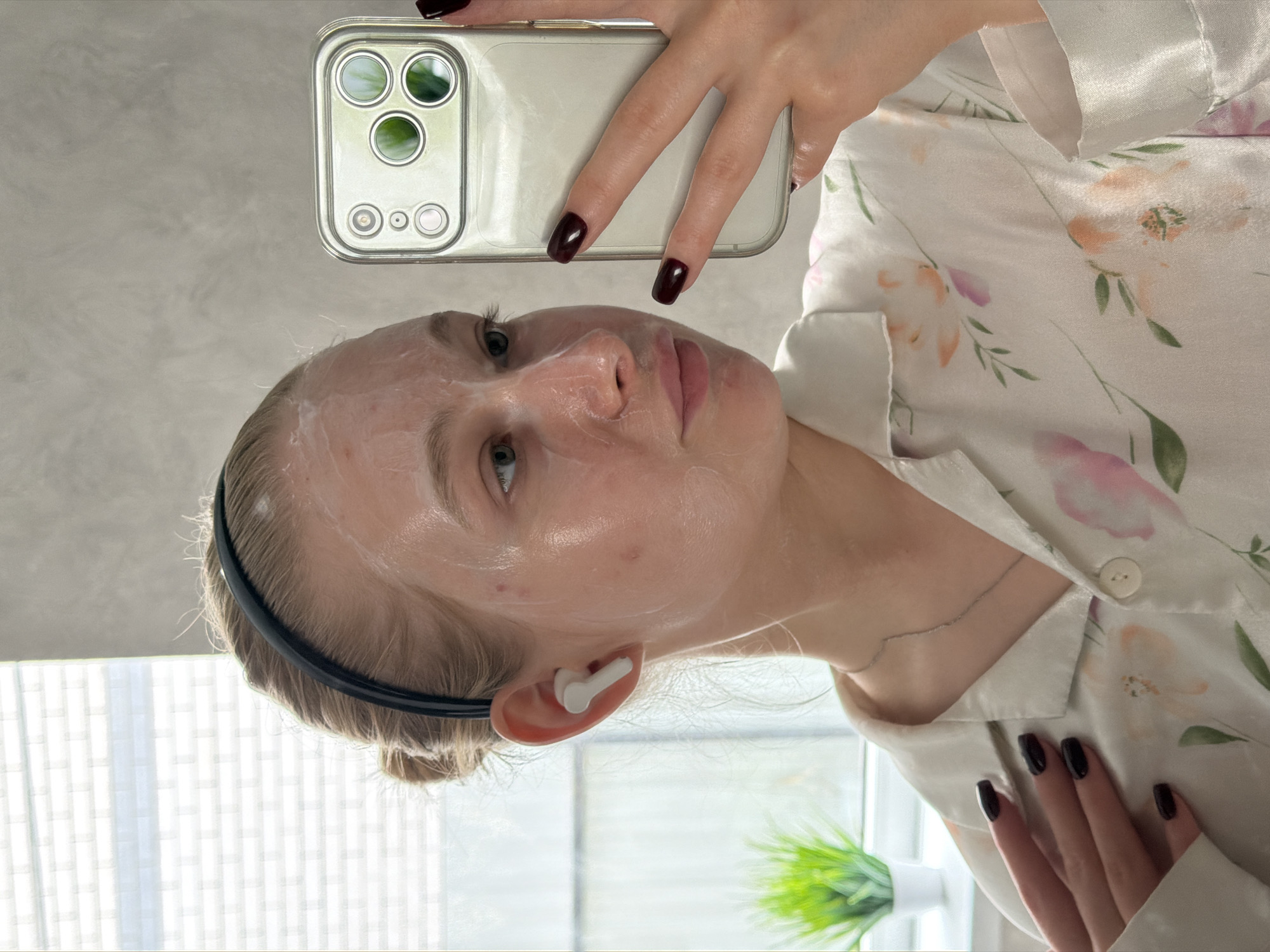 A young woman with fair skin and light blonde hair, held back by a black headband, takes a selfie using a modern smartphone with three prominent lenses. She is wearing a floral-patterned pajama top and has a white facial mask applied to her face. A small white wireless earbud is visible in her left ear. Her nails are painted a dark burgundy color.
The background suggests an indoor setting, with a window and a small green potted plant visible. The location cannot be determined to be Rossiyskiy, Russia with the given information.