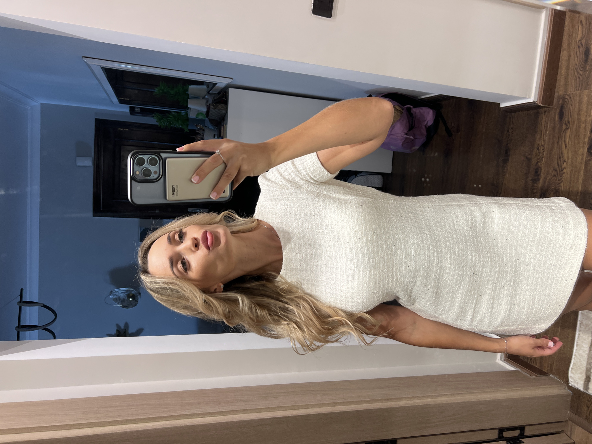 A blonde woman in a white textured dress takes a selfie in a hallway with a mirrored wall. She is pouting her lips and holding up her phone to capture her reflection. The hallway has wooden floors and doors. In the reflection, other items are visible, including a dresser with decorative plants and a doorway leading to another room. The image is clear and well-lit, allowing for detailed observation of the woman and her surroundings. The specific location of Sharm El Sheikh, Egypt, is not indicated in the image itself.