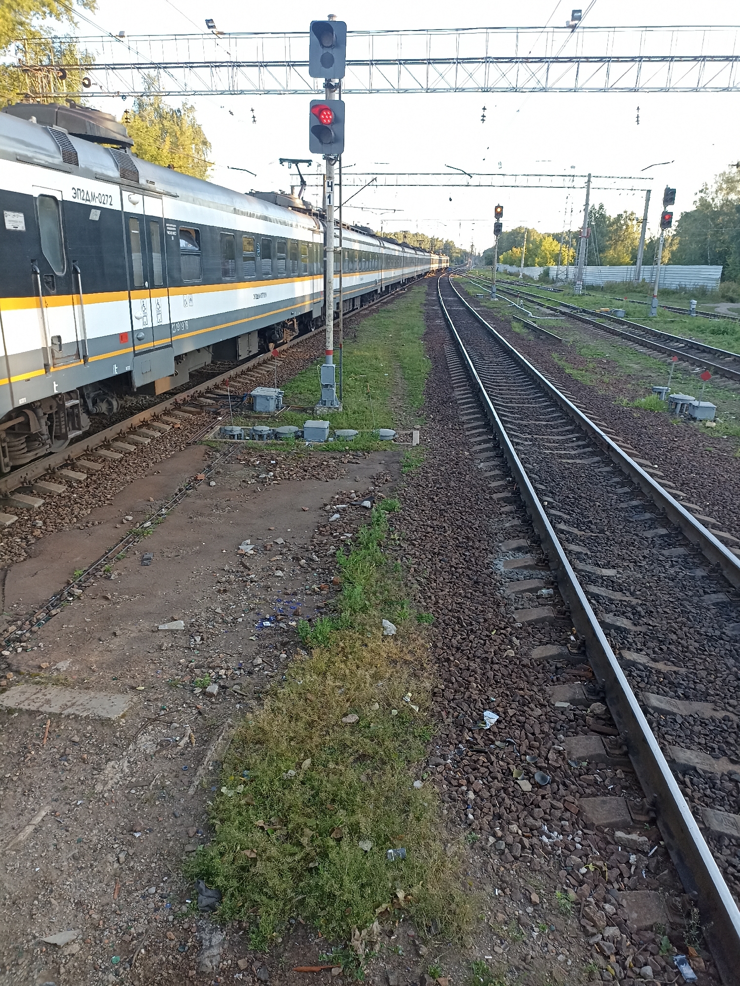 The image shows a train on a railway track in Chekhov, Russia. The train is dark gray and white with yellow stripes and the number "ЭП2ДМ-0272" visible on its side. A railway signal with a red light is visible to the right of the train. The surrounding environment consists of more railway tracks, gravel, grass, and trees in the background. The sky is overcast. The scene appears to be an outdoor railway station or yard.