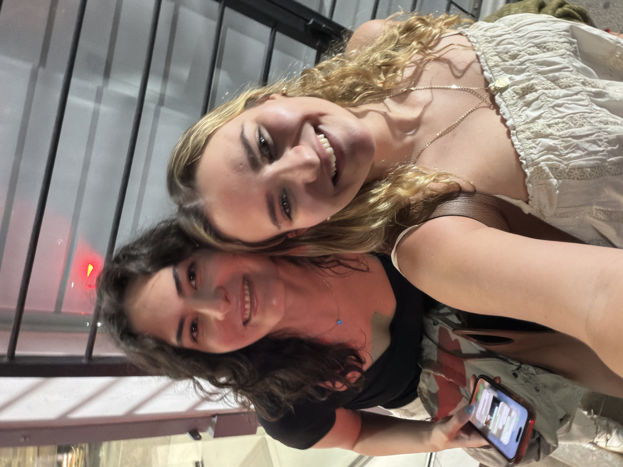 Two young women are smiling at the camera in a selfie-style photograph. The woman on the top has blonde, curly hair and is wearing a white, textured top with a delicate gold necklace. The woman on the bottom has dark, wavy hair and is wearing a black top with a blue necklace. She is holding a smartphone in her hand.
The background appears to be an exterior setting with a metal grate or bars visible, and there's a blurred reflection suggesting glass or a window. A red light is also visible in the background. Based on additional metadata not visible in this image alone, this photo was taken in Puebla, Mexico.