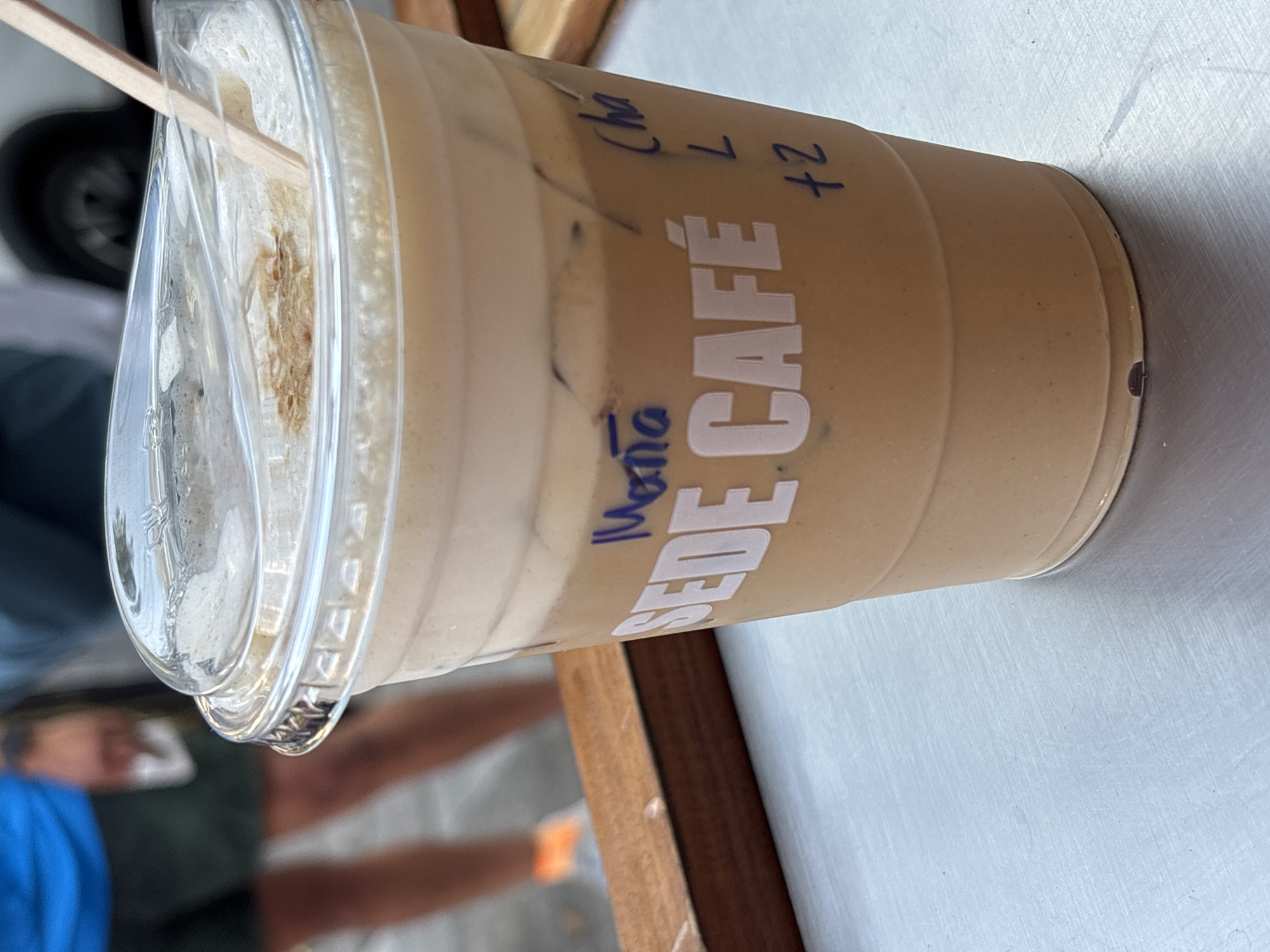 An eye-level shot captures an iced beverage in a clear plastic cup, set on a wooden surface, with a blurred background suggesting an outdoor or semi-outdoor setting in Mexico City, Mexico.
The main subject is a tall plastic cup filled with a creamy, light brown iced drink, likely an iced coffee or chai latte, topped with a layer of foam and a wooden stirrer inserted through the lid. Ice cubes are visible within the drink. The cup features white branding with the words "SEDE CAFÉ" printed vertically. Additionally, there are handwritten notes in blue marker on the cup: "Mona" (likely a customer's name) is written vertically, and "Cha L +2" appears horizontally, possibly indicating "Chai Latte, Large, plus 2 modifications or extra ingredients."
The cup rests on a warm-toned wooden surface, possibly a table or counter, with a lighter, possibly white or light gray, surface visible next to it. The background is softly blurred but shows hints of activity. A person wearing a blue shirt is visible on the left, and another blurred figure with an orange band on their arm can be seen further back. A dark car wheel is partially visible in the upper left background, reinforcing the idea of a street or outdoor cafe environment.
The lighting is bright, suggesting it is daytime. The presence of an iced drink further implies warm weather. The overall scene conveys a casual, on-the-go atmosphere, typical of someone enjoying a refreshing beverage at a cafe or while out and about.