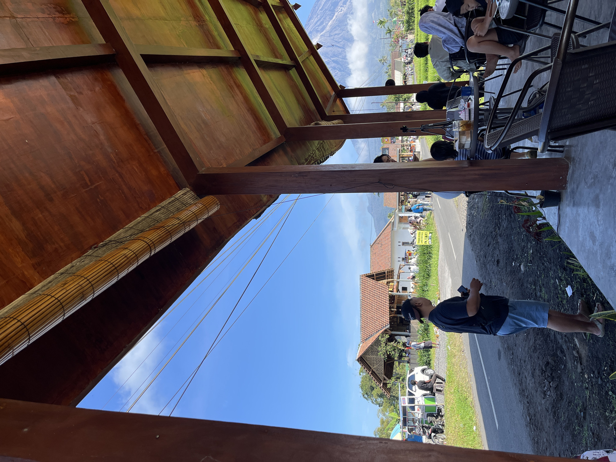 The image depicts an outdoor cafe with a wooden roof structure in the foreground, overlooking a road, some buildings, and a large mountain in the background, likely Mount Merapi, given the location in Oro-oro Ombo, Indonesia. Several people are seated at tables in the cafe, some of whom are engaged in conversation or looking towards the surroundings. A man in a black t-shirt and blue shorts stands near the edge of the cafe, looking towards the road. Beyond the road, a white car and a motorcycle are parked, and other people are walking around. The buildings have red tile roofs, and the landscape is generally green, with some vegetation and rice paddies. The sky is clear and blue. The overall atmosphere suggests a relaxed setting with a scenic view.