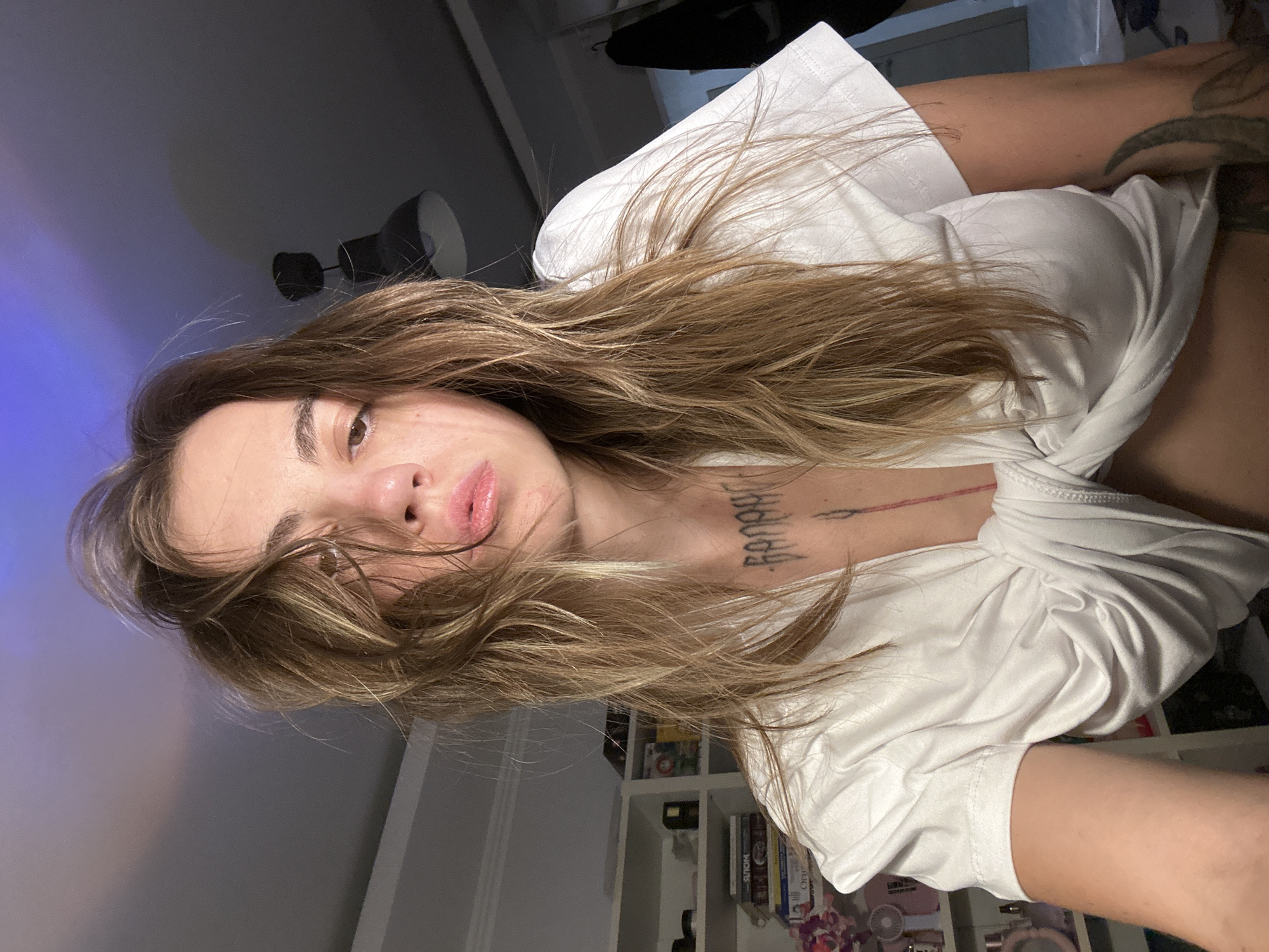 A woman with long, light brown, messy hair, wearing a white t-shirt knotted at the front, is pictured from the chest up. She has tattoos on her chest and arm. The background shows a shelf filled with books and other items, a ceiling light fixture, and a dark doorway. The lighting suggests an indoor setting, possibly a bedroom or living area, with a blue light illuminating the ceiling. The location is not specified in the image or provided context.