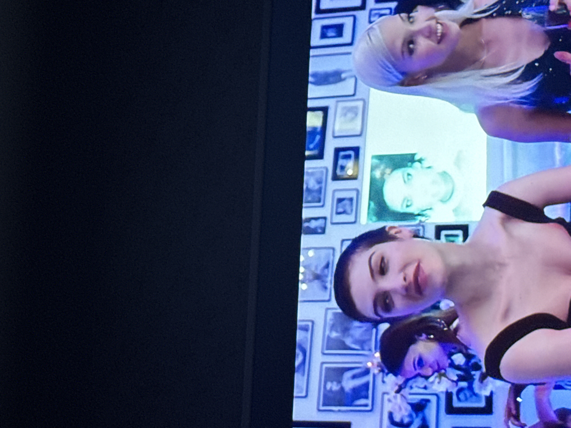 The image shows a portion of a television screen displaying a scene with at least two women in formal attire. The woman in the foreground has short, dark hair and is wearing a black dress. A second woman is behind her, smiling, with long, light blonde hair and a dark, speckled dress. Behind the women is a wall covered with framed black and white photographs. The left side of the image is a dark, blank area which appears to be the edge of the television screen. There is no indication this image was taken in Mytishchi, Russia.