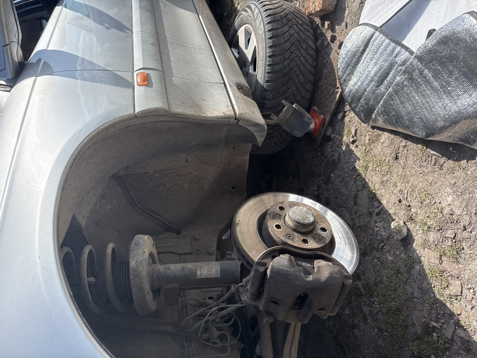The scene depicts a silver-colored vehicle undergoing maintenance outdoors in Asenovo, Bulgaria. The front left wheel of the vehicle has been removed, exposing the brake rotor, caliper, and suspension assembly, which includes a spring and strut. The vehicle is elevated by a red hydraulic jack positioned beneath the frame, resting on a wooden plank, with a black support component atop the jack. Directly beneath the side of the vehicle, a removed wheel with a winter tire rests on the ground. The ground surface is composed of dry, uneven dirt with sparse patches of dry grass and scattered small rocks. In the foreground, a folded piece of reflective material, possibly a car sunshade or insulation, lies on the ground. A single red brick is visible near the removed tire. The scene is illuminated by strong sunlight, casting distinct shadows on the ground, indicating clear weather. No individuals are visible within the frame.