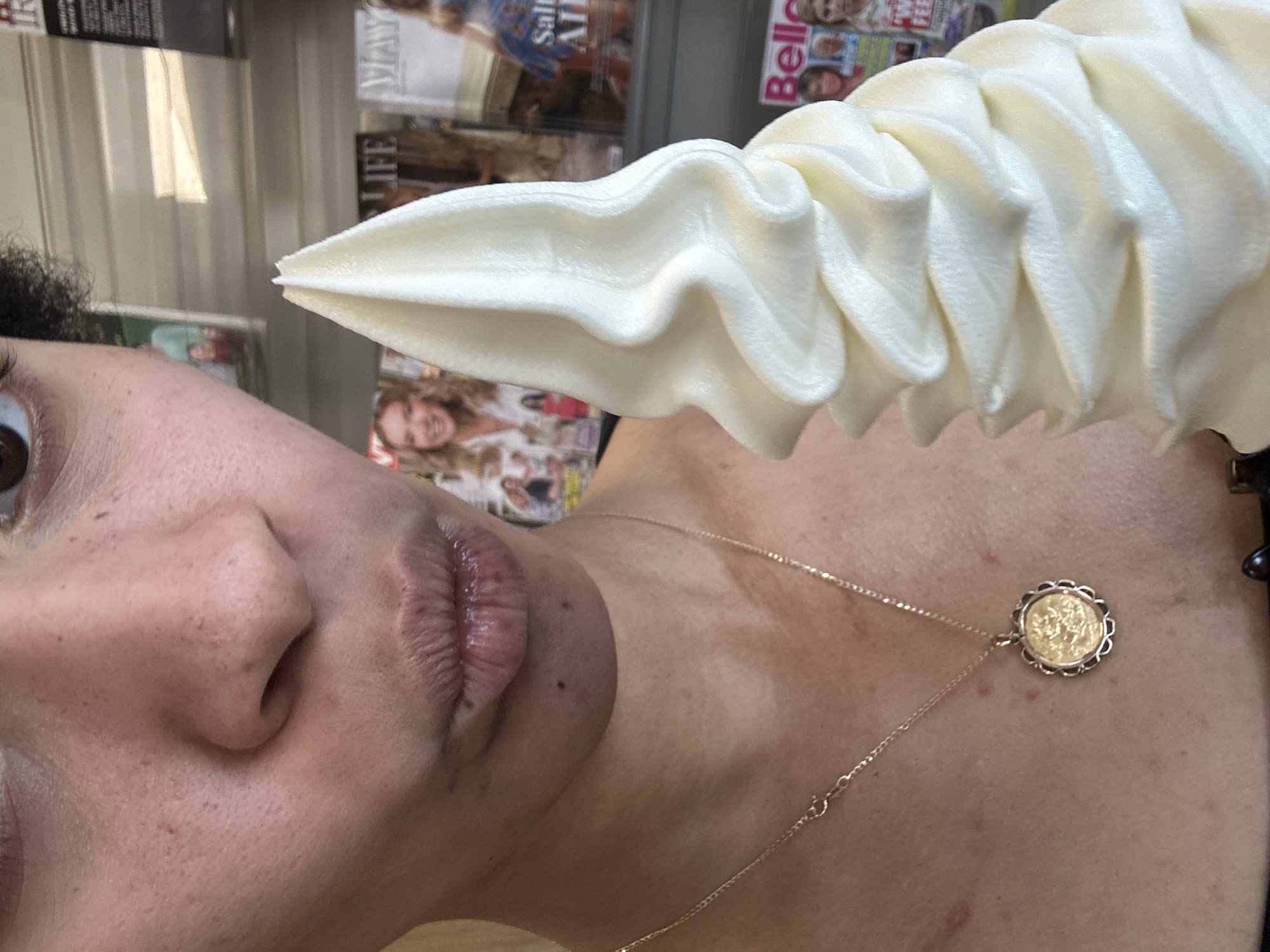 A close-up, vertically oriented, medium shot captures the left side of a person's face, focusing on their nose and lips, along with a portion of their neck and chest adorned with a gold necklace. The person, with curly dark hair partially visible on the left, appears to be looking away from the camera, with their lips slightly parted and glossy. The skin shows some natural texture and a few freckles.
Dominating the upper portion of the frame is a large, swirled soft-serve ice cream cone, its creamy white texture catching the light. The background is blurred, but suggests an indoor environment, possibly a store or market, with shelves of magazines visible. The magazine titles are partially obscured, but "LIFE" and "MAY" are discernible on some. Another magazine has "Belle" printed on it.
The lighting is bright, suggesting daytime. The scene seems casual, with the individual enjoying the ice cream, possibly as a treat or snack. The overall impression is one of a candid, everyday moment. There are no overt emotional expressions, but the focus on the food suggests enjoyment. The context of Westport, Ireland, is provided, though no specific landmarks are visible within the frame.
