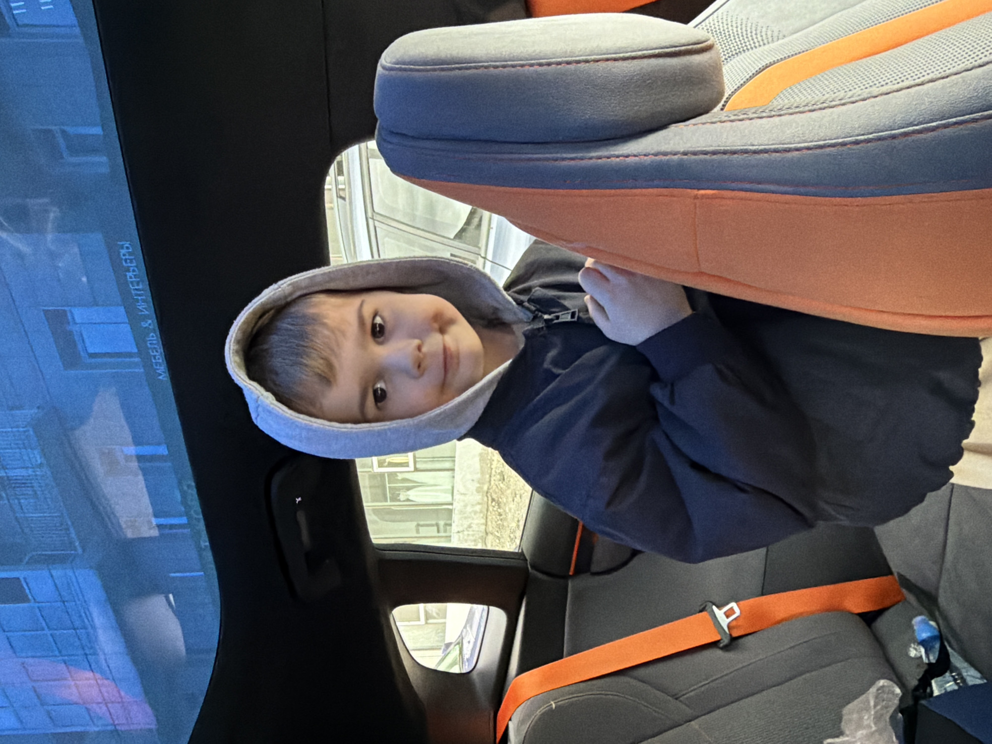 A boy wearing a gray hooded jacket sits in the backseat of a car. The car has gray and orange seats with matching orange seatbelts. Outside the window, the buildings suggest a location in the city of Krasnoyarsk, Russia. A screen or monitor is visible, displaying the words "мебель & интерьеры" (furniture & interiors).