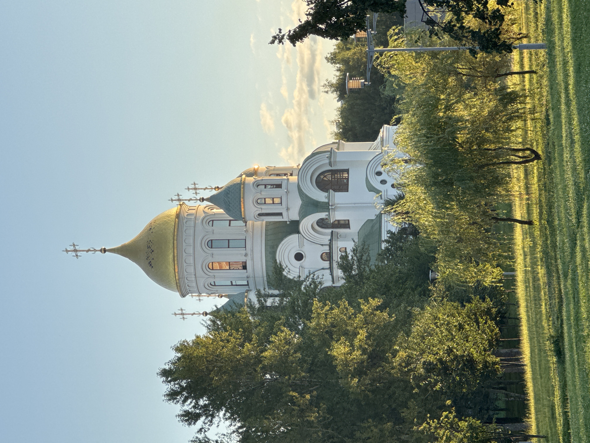 The image shows the Church of the Resurrection at Patriot Park in Moscow, Russia, at sunset. The white church has a golden dome and green accents. It is surrounded by lush green trees and a grassy lawn, with the setting sun casting long shadows. The sky is a clear blue with a few wispy clouds. There are no people visible in the image.