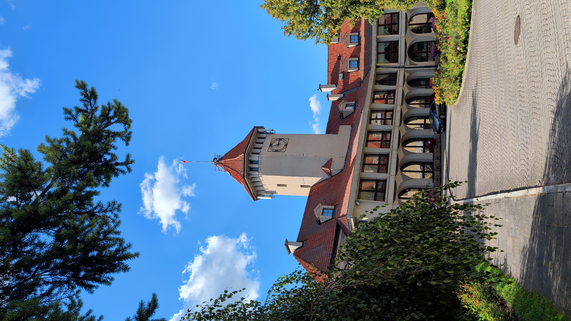 The image shows the exterior of a hotel in Pułtusk, Poland. The building has a prominent clock tower with a red tiled roof, and a long, lower section also with red tiled roofs and numerous windows. A paved driveway leads up to the hotel entrance, which is lined with arched windows. Flower beds with colorful blooms are visible in front of the hotel. A dark-colored car is parked on the driveway. Green trees and branches frame the scene, with a bright blue sky and scattered clouds above. The scene appears to be a sunny day.