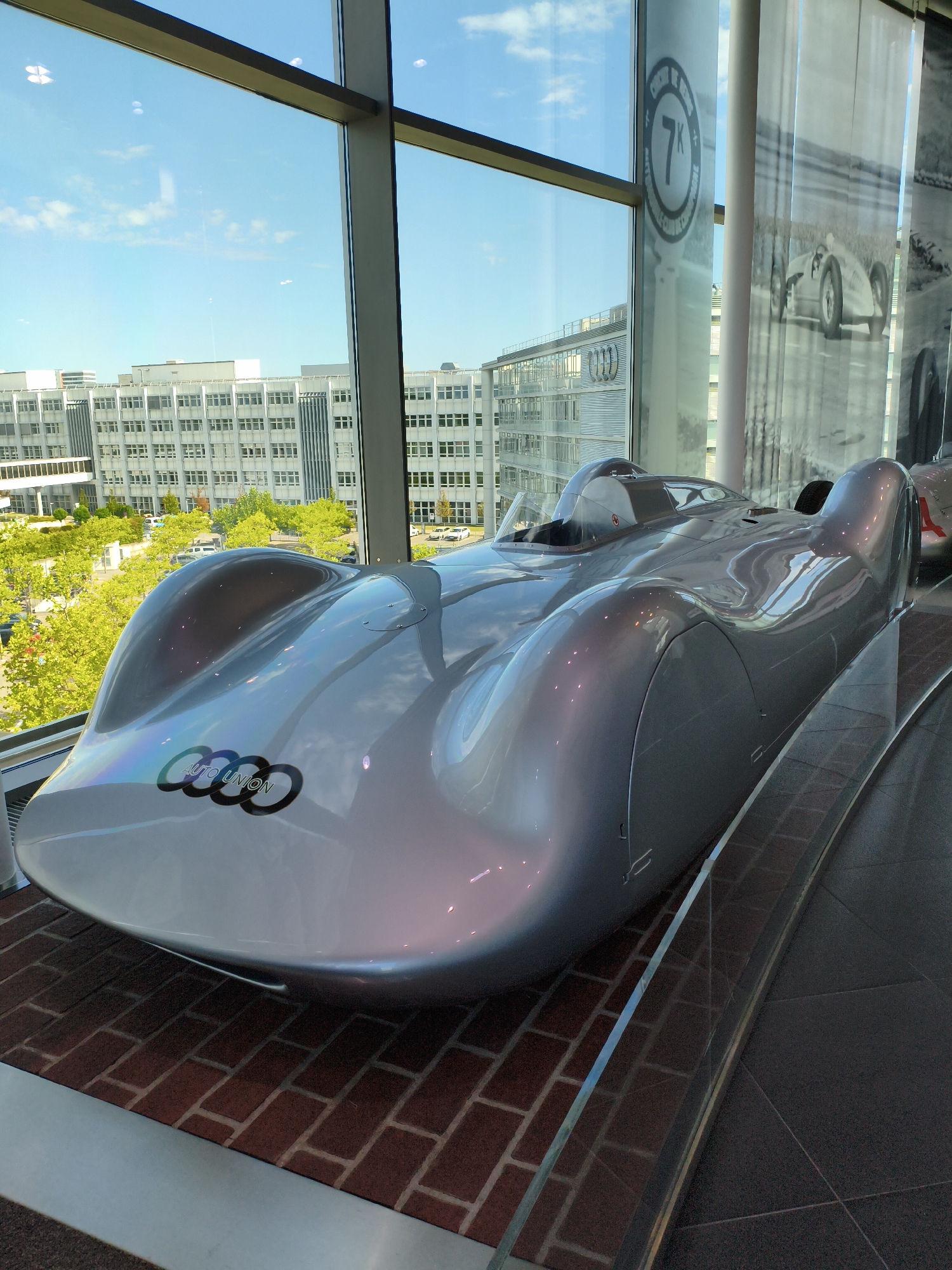 The image shows an Audi Type C Streamliner, a silver-colored vintage race car, on display in a museum. The car is positioned diagonally on a brick floor, with a glass barrier in front of it. Behind the car, through a large window, are modern office buildings and trees, suggesting an urban environment. The window also reflects the sky and clouds. To the right of the car, there are large printed banners or curtains depicting historical race cars and figures. The overall scene appears to be within the Audi Forum Ingolstadt, a museum and brand experience center located in Ingolstadt, Germany. There are no people visible in the image.