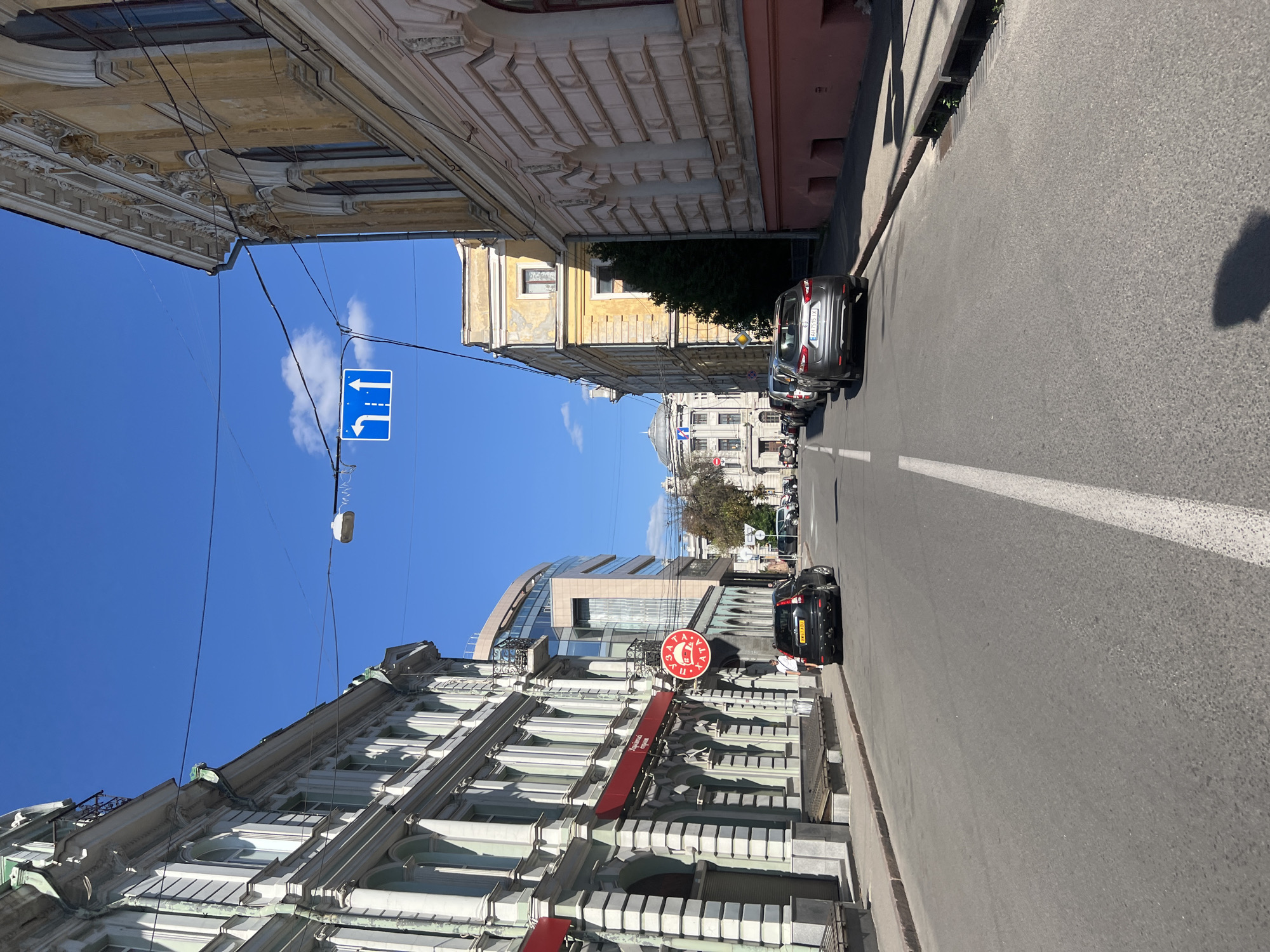The image shows a street scene in Kharkiv, Ukraine, on a sunny day with a clear blue sky. The street is paved and has white lane markings. Several cars are parked on the right side of the street and one car is driving down the street. On the left side of the street, there are buildings with ornate architecture. One building has a red awning. There is a traffic sign overhead indicating that traffic can go straight or turn left. In the distance, a large, light-colored building with a domed roof is visible. The overall impression is of a city street with historical buildings.
