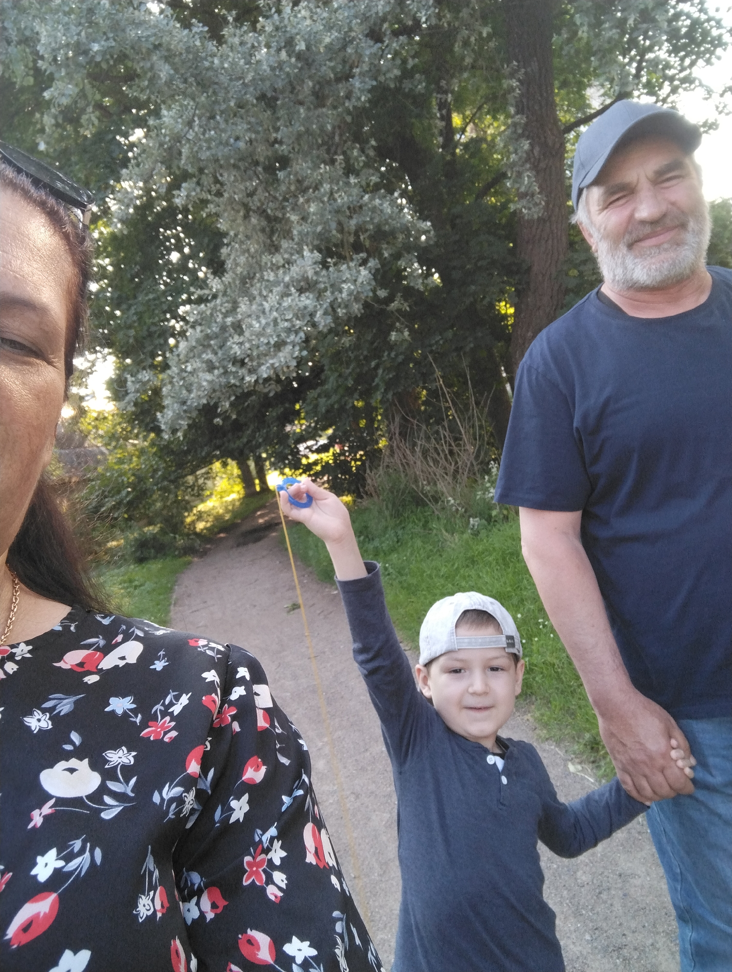 The image depicts a family outing in what appears to be a park or wooded area with a dirt path. A woman, partially visible on the left, is wearing a black floral-patterned dress and a gold chain necklace. To her right, a young boy wearing a gray baseball cap and a dark long-sleeved shirt is holding a blue toy with a yellow string. The boy is holding hands with an adult man on his right, who is wearing a dark blue t-shirt and a blue baseball cap. The man has a gray beard and is smiling at the camera. Behind the people, there are trees and foliage, suggesting an outdoor, natural setting. The lighting indicates it is daytime, possibly late afternoon. The presence of a path suggests a location suitable for walking or recreation. While the exact location is not specified, it is described as being in the city of Kingisepp, Russia.