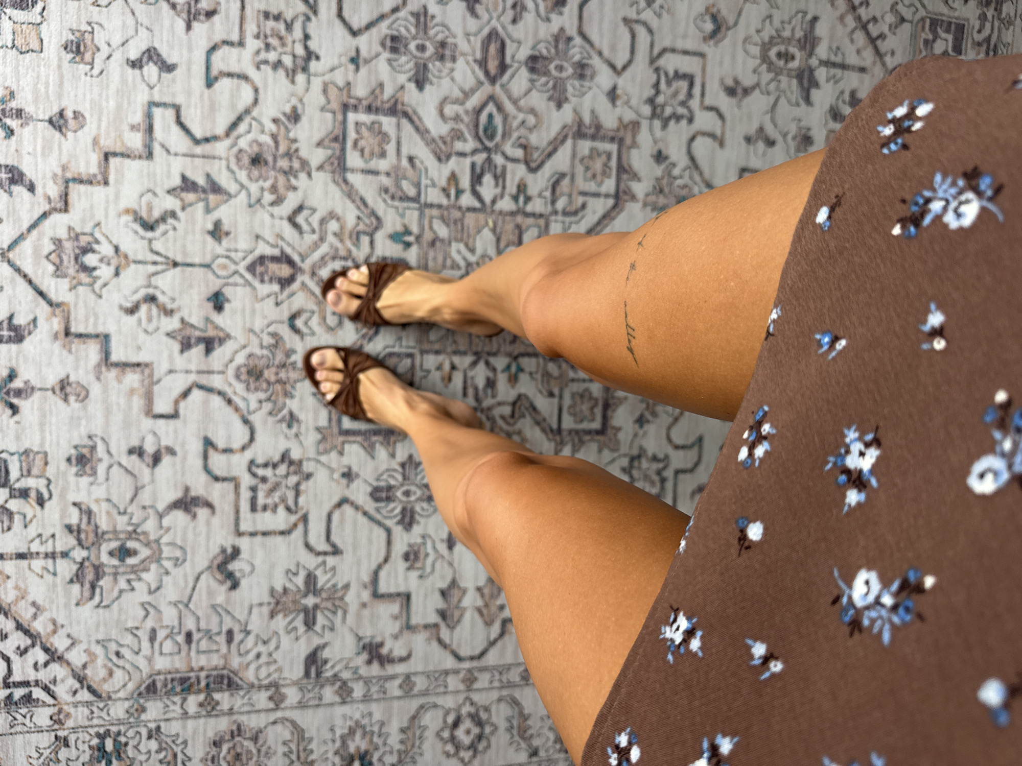 The image depicts a person from an elevated, overhead perspective, revealing only their legs and feet. They are standing on a patterned rug with a predominantly grey, blue, and beige floral and geometric design. The person wears brown sandals with a strap detail over the toes. A portion of a brown dress with small blue and white floral patterns is visible on the right side of the frame. A faint tattoo is visible on the right thigh. The setting, Tibubeneng, Indonesia, is assumed from the prompt.