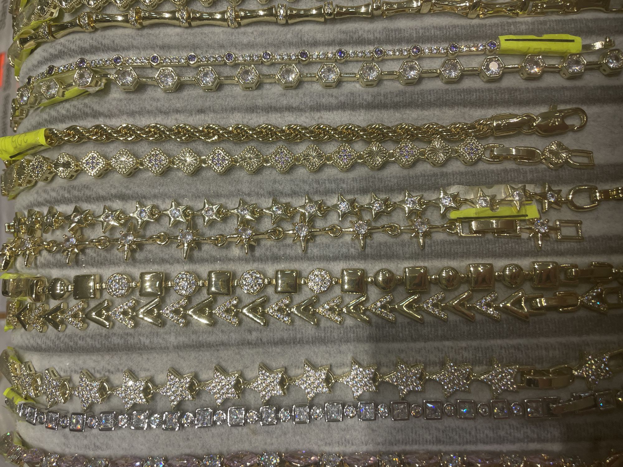 This image displays a close-up, top-down view of several gold and silver bracelets laid out on a grey, textured surface. The bracelets are of various designs, featuring chains with different charms and embellishments, including stars, geometric shapes, and what appear to be small gemstones or cubic zirconia. Some bracelets are predominantly silver with clear stones, while others are gold with clear and purple stones. The overall impression is of a jewelry display. There is no information to suggest the image's location in Yunus Emre, Turkey. The image shows only the bracelets and the surface they are resting on, with no people or actions depicted.