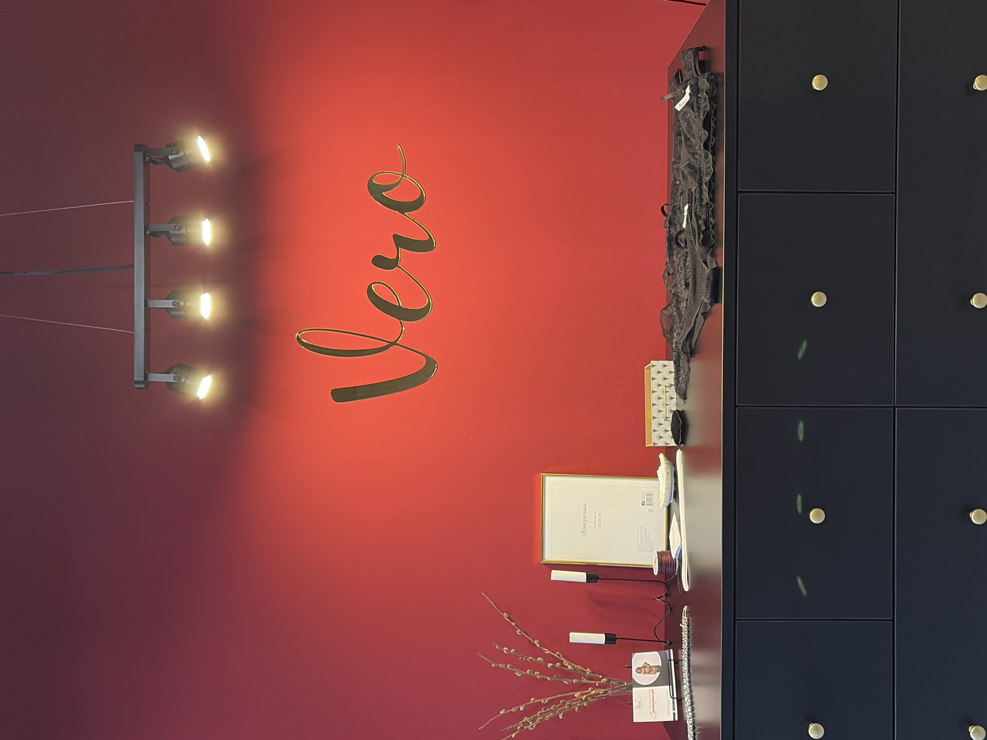 The image shows a close-up interior shot of a retail space, likely a boutique, with a bold red wall as the backdrop. A metallic gold cursive script spelling "Vero" is affixed to the wall. Below the script, a dark countertop displays various items. To the left, a small bouquet of pussy willow branches in a glass vase sits next to a framed certificate. Two black candlesticks, one with a white candle, are also present. To the right of the frame, there's a decorative box with a geometric pattern and a piece of dark lace lingerie draped over the countertop. The countertop is mounted on a dark dresser with multiple drawers, each featuring a single round brass knob. The overall impression is one of a display area within a shop. Based on the context of "Vero Moda" stores, this could be a Vero Moda store in Chaikovsky, Russia, as Vero Moda is a popular clothing brand with many international locations.