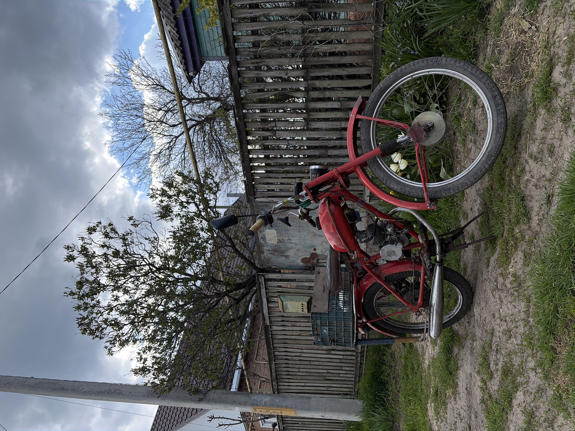 A red moped stands parked on a dirt path in a rural or semi-rural location. The moped is positioned in front of a weathered wooden fence and several outbuildings. A leafless tree extends into the sky above the moped. The sky is partly cloudy, with a mix of blue sky and dark storm clouds. The surrounding area has patches of green grass and weeds. The location appears to be in the city of Unknown, Unknown.
