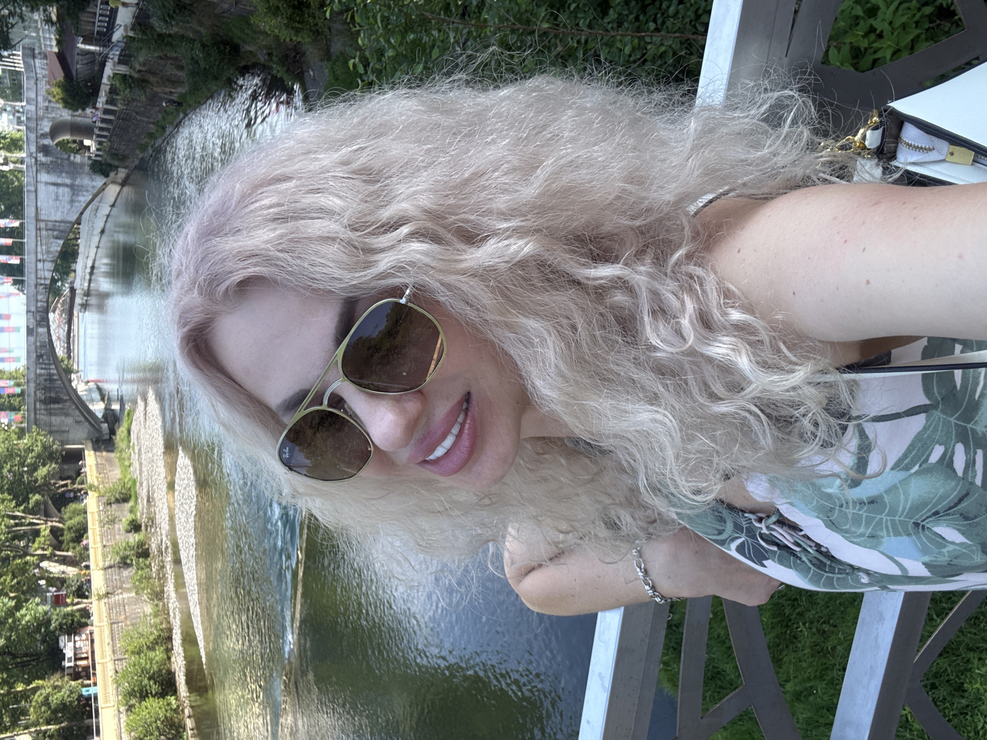 The image is a selfie taken by a woman with long, wavy blonde hair. She is wearing sunglasses and a light-colored top with a green leaf pattern. She is smiling and appears to be outdoors. In the background, there is a body of water with a stone embankment and lush green trees. A bridge can be seen in the distance. The scene suggests a sunny day in a scenic, possibly urban, location. The woman's pose and smile convey a relaxed and happy mood. The details in the background, such as the architecture of the bridge and the style of the waterfront, do not provide specific cues to distinguish it from other locations globally, though the prompt context specifies Sochi, Russia. No visible text is present in the image.