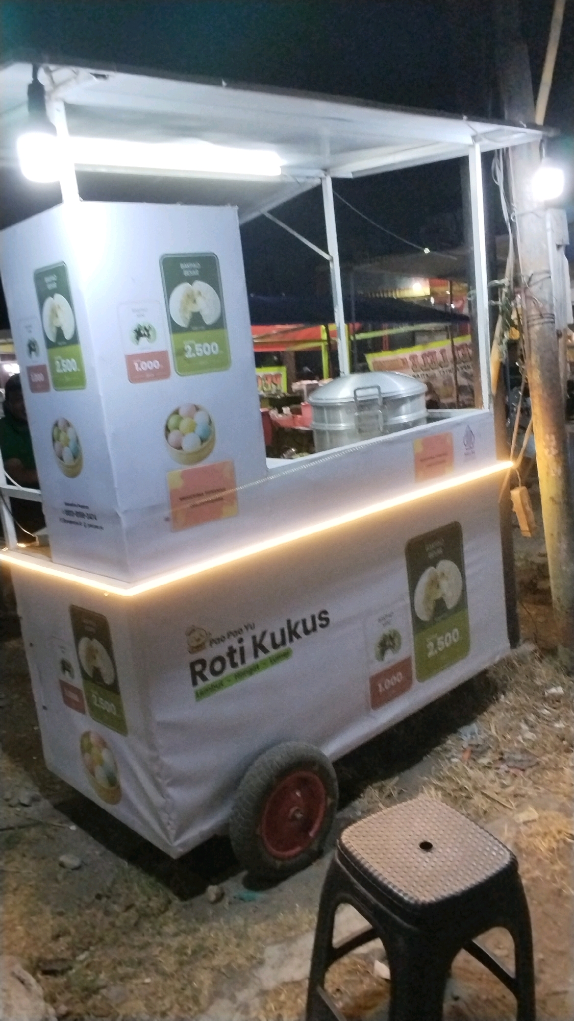 This is a medium-shot, eye-level, outdoor, nighttime photograph of a food cart in Canggu, Indonesia. The cart is white with a light-up trim around the bottom and a white canvas roof supported by metal poles. The cart is selling "Roti Kukus," or steamed buns, which are advertised on the side with colorful images of the buns and prices such as 2,500 and 1,000. A large metal steamer sits on the cart. To the right of the cart, a black plastic stool is visible. The cart is parked on a dirt and grass surface, with debris and dry grass scattered around. In the background, there are blurry lights and structures, suggesting a commercial or street food area. The overall lighting is dim, with the cart and its advertisements being the primary illuminated subjects.