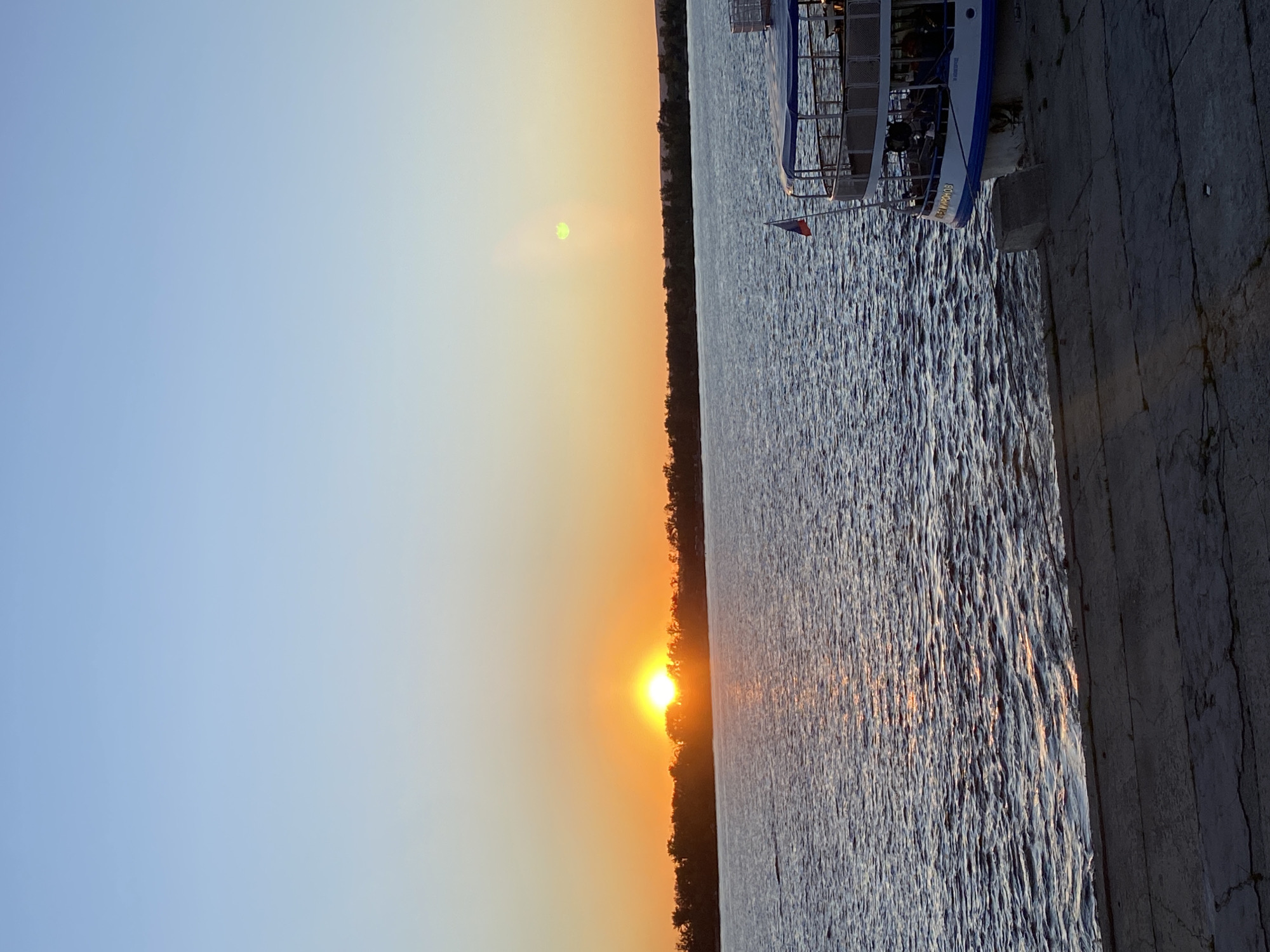 The image depicts a sunset over a body of water, with a boat docked along a concrete embankment. The sky transitions from a clear blue at the top to a warm orange hue near the horizon, where the sun is setting. The sun itself is a bright, glowing orb, partially obscured by a dark line of trees along the distant shore. Its light reflects on the rippling surface of the water, creating streaks of golden and orange light.
A multi-decked boat, painted white with blue trim, is tied to the embankment. Several people are visible on the boat's decks. A small flag, appearing to be the Russian flag, is attached to the boat's railing. The embankment is made of rough, gray concrete, showing signs of wear and age with visible cracks and texture.
The overall mood of the image is peaceful and serene, characteristic of a sunset. The time of day is clearly evening, as indicated by the setting sun. The weather appears to be clear, with no clouds visible in the sky. The scene suggests a waterfront location, possibly a river or a lake, where recreational boating takes place. The name "В. МАЯКОВСКИЙ" is visible on the side of the boat.