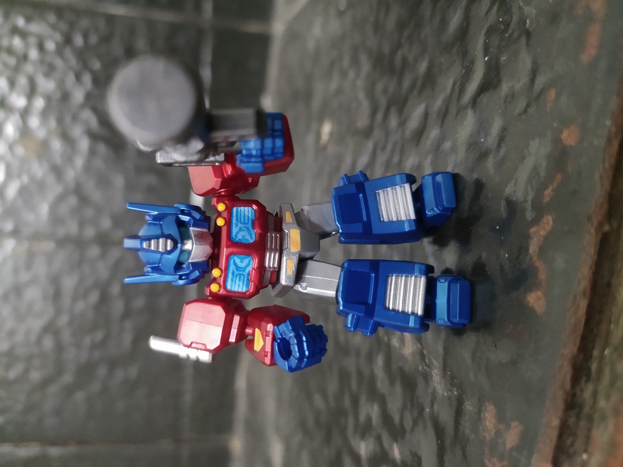 A small, blue and red Optimus Prime action figure stands on a dark, reflective surface. The figure is in a standing pose, with its right arm raised and holding a gray weapon. The background is a textured, dark gray surface. There is no indication of the city of Ngantru, Indonesia, in the image.
