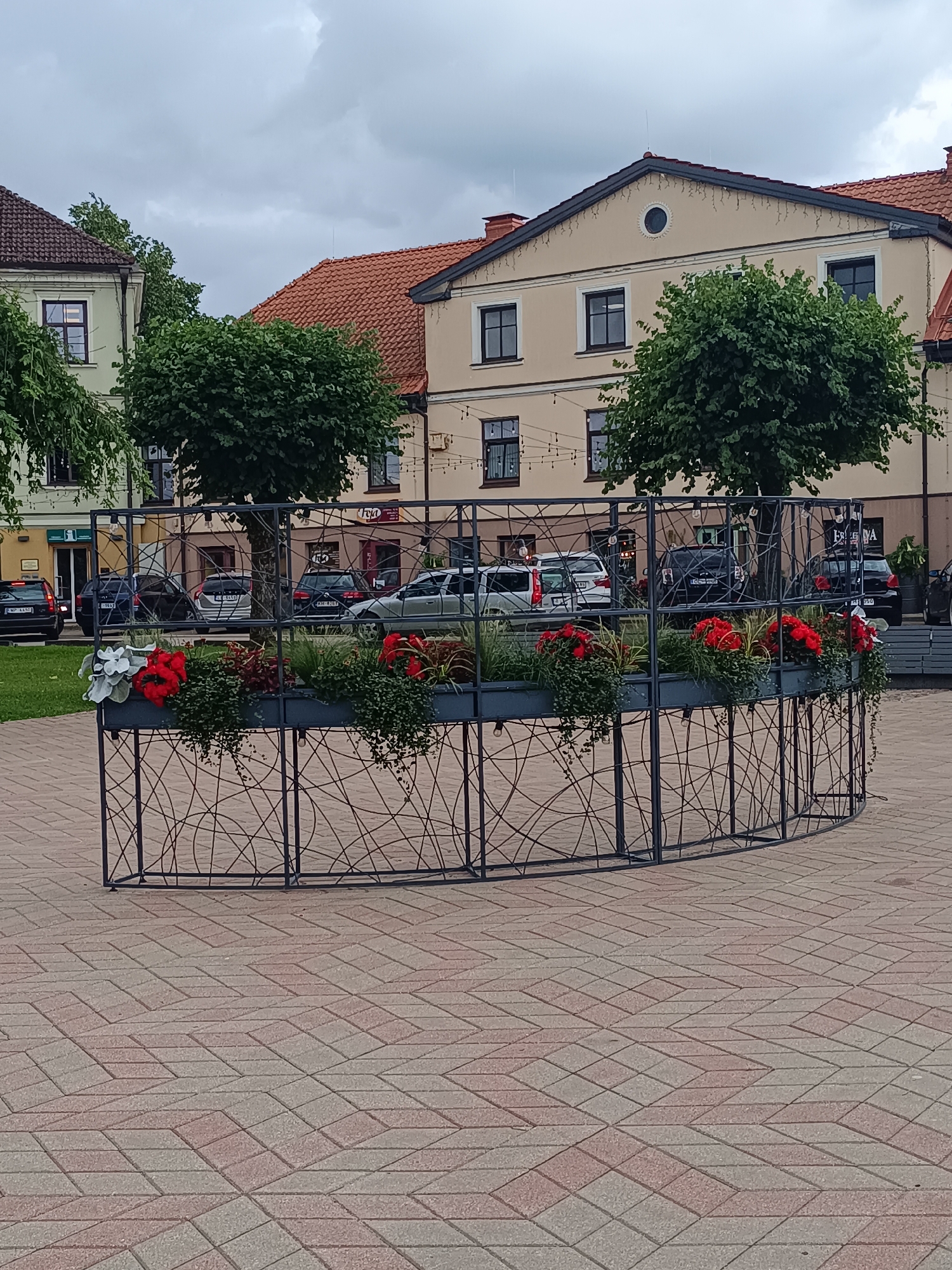 In Saldus, Latvia, a paved public square features a large, curved planter box filled with red and white flowers and greenery. The planter is enclosed by a decorative black metal fence with a swirling pattern. Behind the planter, several cars are parked in front of buildings with red tiled roofs. Trees with lush green foliage are visible between the planter and the buildings. The sky is overcast with grey clouds. The ground is paved with interlocking bricks in a diamond pattern.