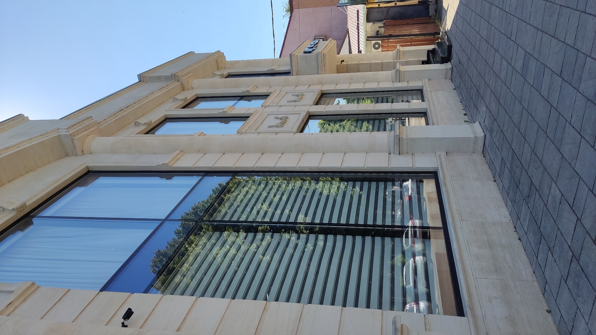 The image shows a modern building with a stone facade and large windows. The building appears to be commercial, possibly an office or retail space, with a stone pavement sidewalk outside. The windows reflect the sky and trees, as well as the interior of the building, showing blinds and some reflections of cars. There is a sign on the building that appears to say "Arno" in Cyrillic. The overall impression is one of a well-maintained and upscale establishment. The sky is clear and blue, suggesting a sunny day. The time of day appears to be late morning or early afternoon. The location is Krasnodar, Russia.
