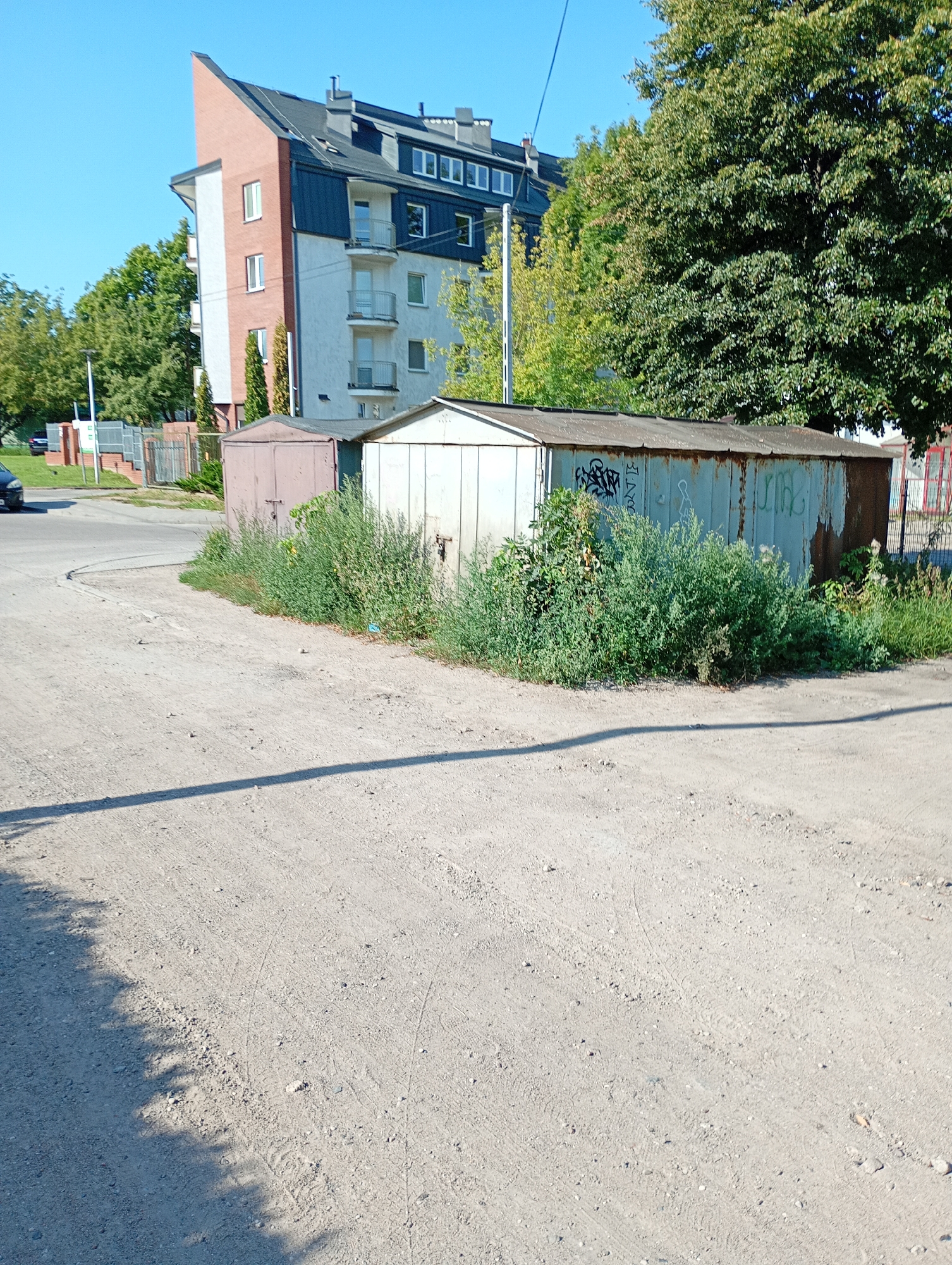 The image shows a street scene in the city of Białystok, Poland, on a sunny day. In the foreground, a dirt path is visible, casting a shadow of trees on the ground. Behind the path, two old, rusty garages stand side-by-side, partially overgrown with green bushes. One garage is painted a dusty pink, while the other is a faded greenish-blue with graffiti on its side. Further in the background, a modern apartment building with white and pink facades and a dark roof dominates the scene, with lush green trees surrounding it. The sky is clear and blue.