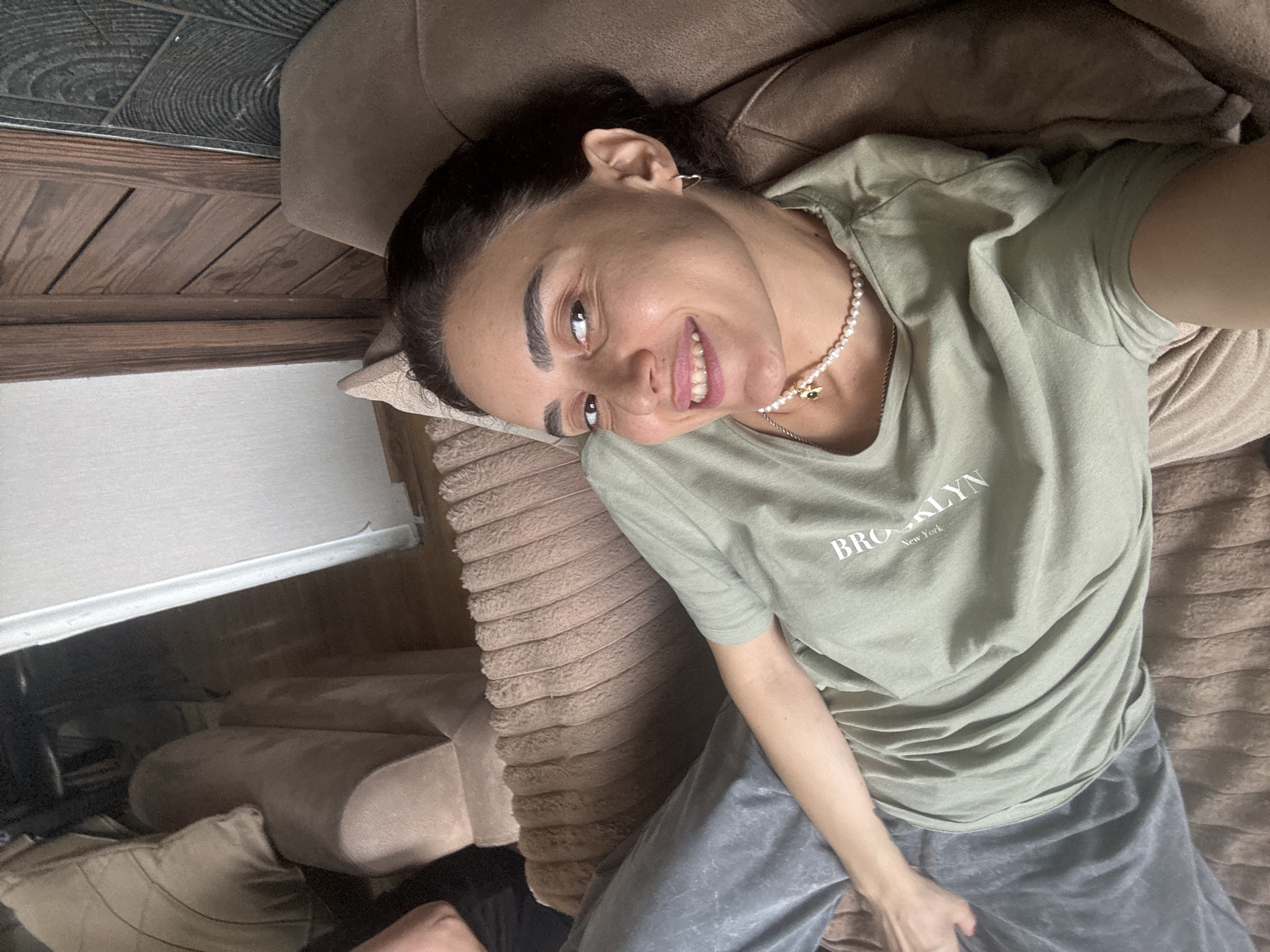 A young woman with dark hair and brown eyes is lying on a couch in an indoor setting. She is wearing a light green t-shirt with "BROOKLYN New York" printed on it and gray sweatpants. A pearl necklace with a small golden pendant is around her neck. Her left arm is resting on her lap, and her right arm is bent, with her hand near her face. The couch appears to be made of a soft, textured fabric. The background shows wooden paneling on a wall and part of another piece of furniture, possibly a chair or another section of the couch. The lighting suggests it is daytime, with some natural light entering the room. The woman is looking towards the camera and smiling. The scene does not provide any specific details that would indicate a location within the city of Tbilisi, Georgia.