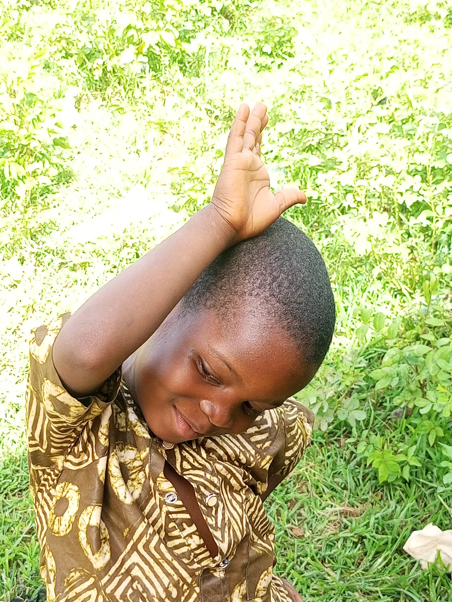 An overhead, medium shot captures a young child wearing a brown and white patterned shirt, looking down with closed eyes and a slight smile. The child's right arm is raised, with the hand resting on the head. The background is a blurred expanse of green grass and foliage, suggesting an outdoor setting. The child appears to be in the city of Tempure, Nigeria, given the context.
