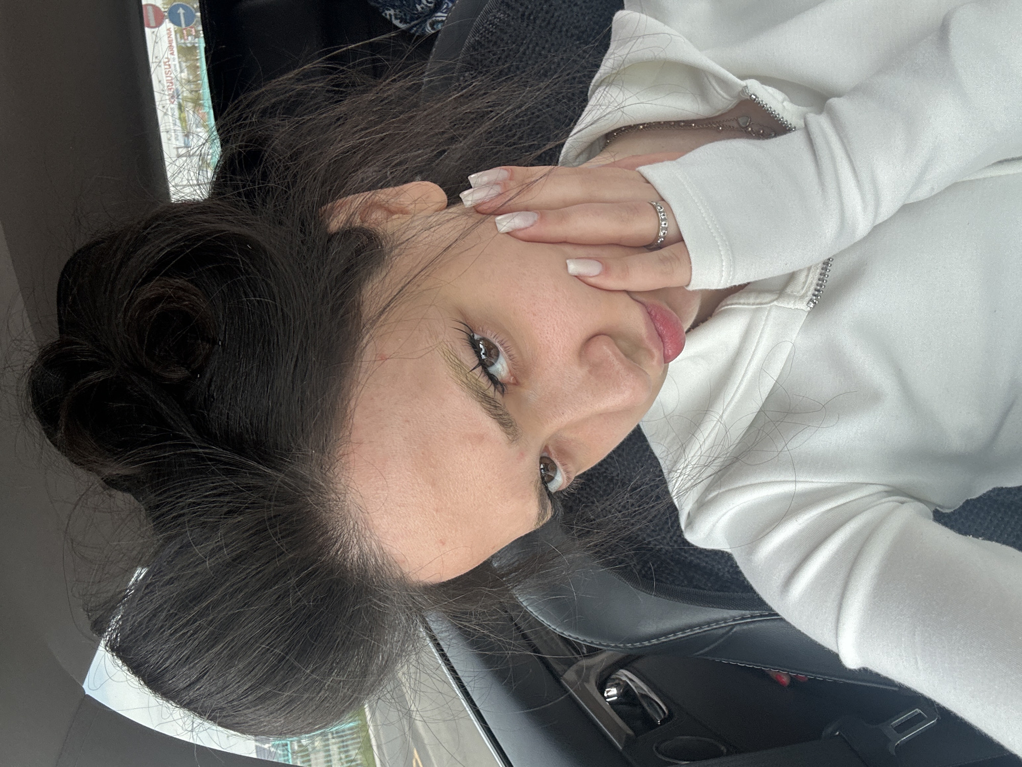A young woman is seated inside a car. She has long, dark hair pulled back into a bun and is wearing a white hooded sweatshirt with the hood up. Her right hand is near her face, with a ring visible on her finger. A silver chain necklace is also visible. The car's interior is dark, and the car appears to be in Yerevan, Armenia. A road sign indicating Yerevan and Armenia is visible through the car window.