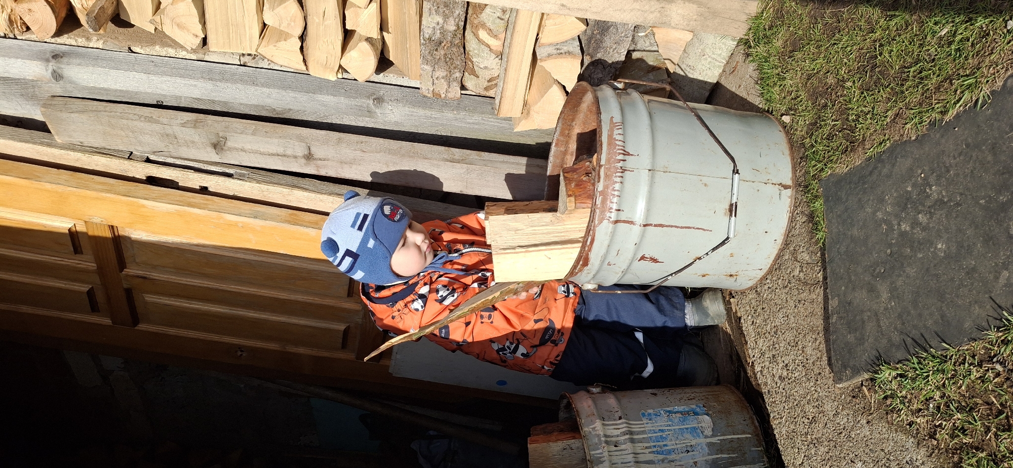 A child wearing an orange panda-patterned jacket and a blue hat with pom-poms is sitting amidst a woodpile, with some wood resting in his lap. Adjacent to him is a light gray, rusty metal barrel laying on its side, containing firewood. To the left are stacked wooden planks, some with bark visible. A segment of a wooden door is also visible on the left. To the right, there's a patch of grass and a black mat. Another barrel is partially visible below the first one. The location is Chaikovsky, Russia.