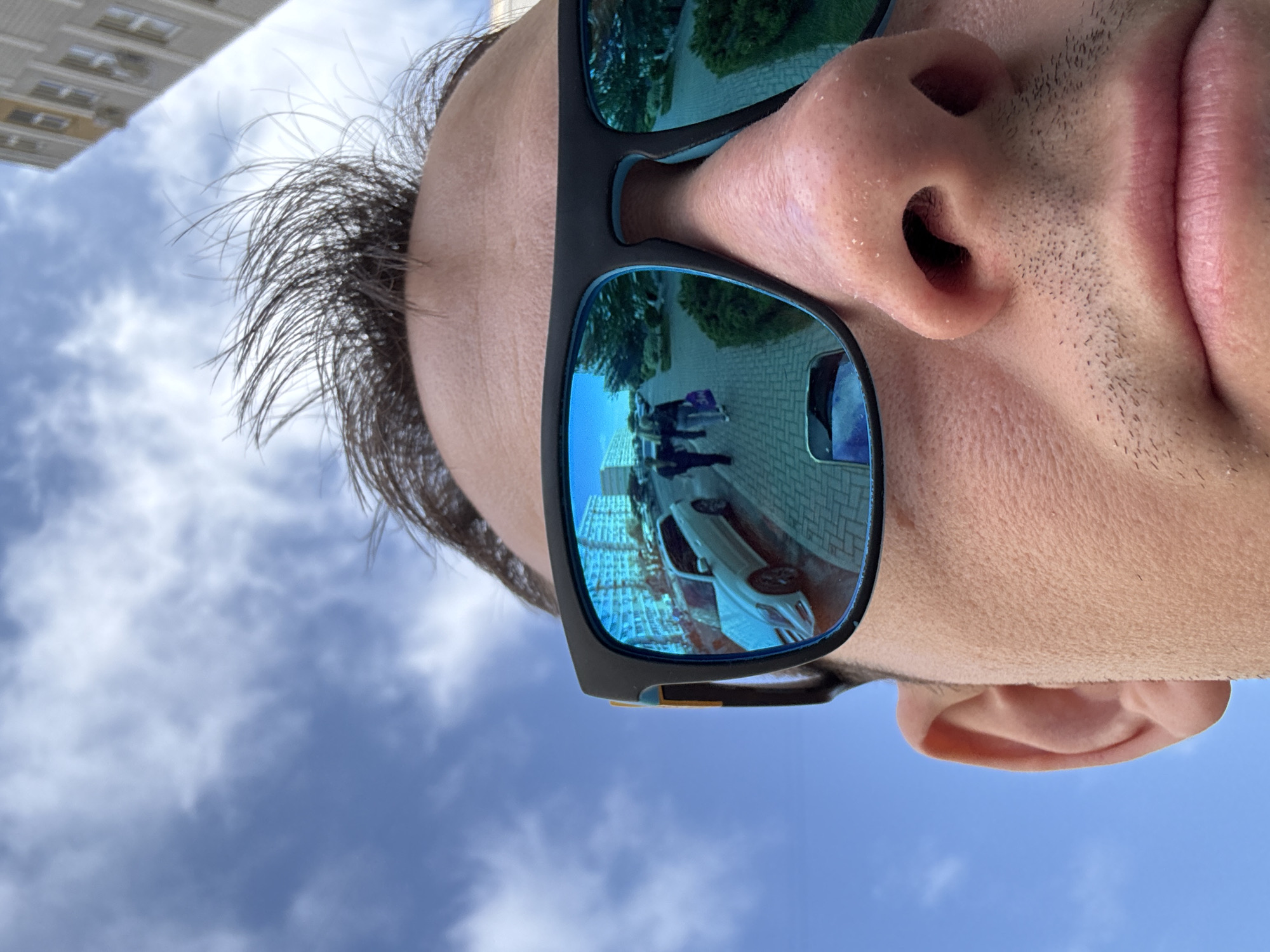 A close-up, low-angle shot of a person's face wearing sunglasses. The reflection in the sunglasses shows a street scene with a white car, people walking, and a building. The building in the reflection is consistent with the architectural style found in the city of Krasnodar, Russia. The sky is visible in the background.