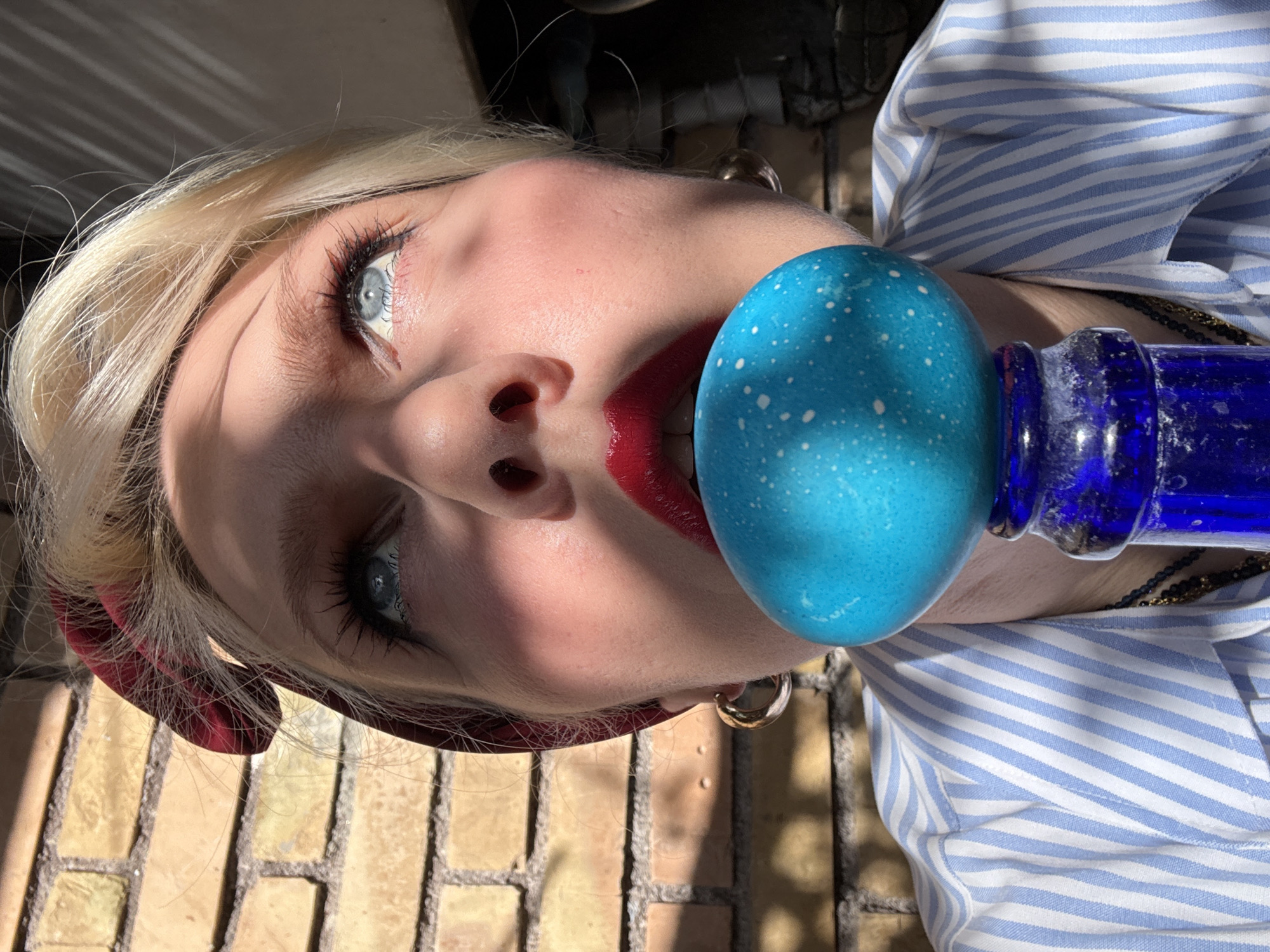 A woman is photographed with a blue, speckled egg and a blue glass bottle. The woman has blonde hair, blue eyes, and wears red lipstick. She has a red headband, gold hoop earrings, and a blue and white striped shirt. A gold and black necklace is also visible. The background includes a brick surface and a dark, indistinct area possibly with pipes or other infrastructure. The location is not identifiable as Rome, Italy, from the given image.