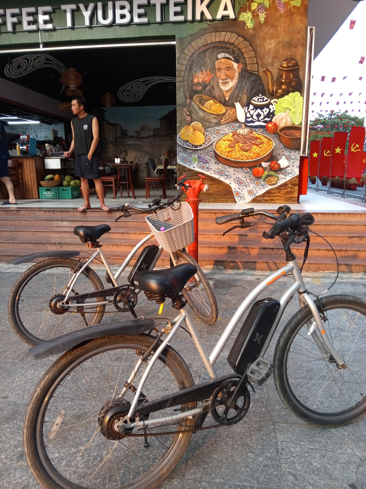 This image presents a street scene in Bắc Nha Trang, Vietnam, captured during the warm light of late afternoon or "golden hour," evidenced by the rich, warm tones and distinct shadows on the steps and ground.
In the foreground, two bicycles are parked on the pavement. The one on the right is a silver e-bike featuring a black battery pack with a "Hurley" logo, while the other, slightly to the left, is a lighter-colored bicycle equipped with a woven front basket.
Behind the bikes, a set of wooden steps leads up to the entrance of a cafe named "COFFEE TYUBETEIKA," clearly visible above the shopfront in large white letters. Inside the cafe, a young man in a black sleeveless shirt and shorts stands by a counter, looking to his left. Beneath the counter, a green "San Miguel Brewery" crate holds various fruits. The cafe's interior shows wooden tables and chairs, with one table displaying the number "7." The back wall features a mural depicting a classic street scene.
To the right of the cafe's entrance, a striking large mural painted on the building's facade depicts an elderly man wearing a traditional "tyubeteika" (skullcap), seated by a hearth, surrounded by traditional Central Asian food like pilaf, fruits, and a teapot, reinforcing the cafe's thematic name. Adjacent to this mural, a row of red flags, each adorned with a yellow star and a hammer-and-sickle symbol, stands prominently, indicative of a communist country like Vietnam. The scene combines everyday street elements with distinct cultural and national symbols, creating a unique visual narrative.