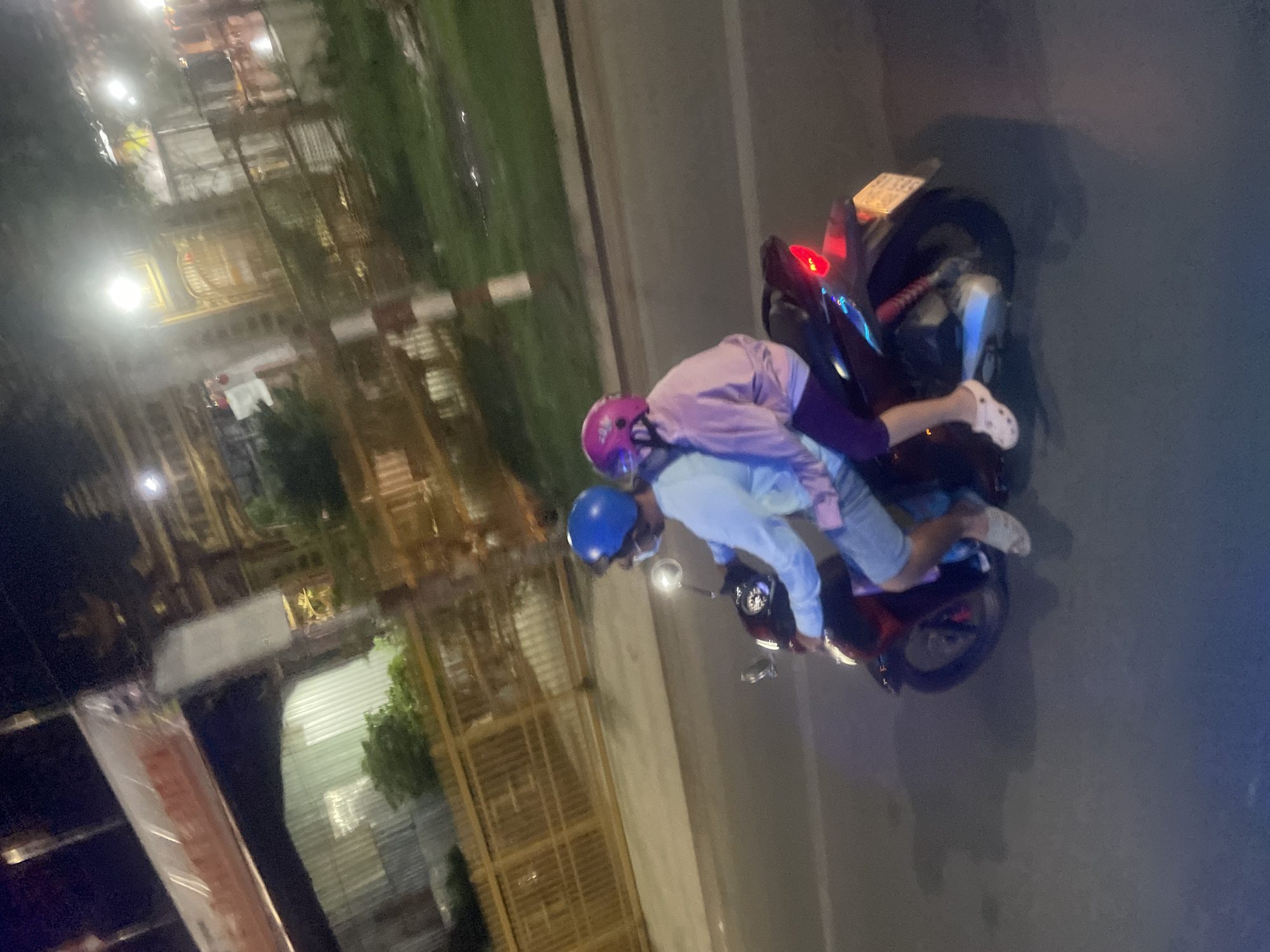 Two individuals, wearing helmets and masks, are riding a motorcycle at night on a paved road. The person in front, wearing a light blue long-sleeved shirt and shorts, is operating the motorcycle. The person behind, wearing a purple long-sleeved shirt and leggings, is a passenger. Both are wearing helmets, one blue and one pink. The motorcycle is dark-colored with a red taillight. The background shows a fence, greenery, streetlights, and buildings, suggesting an urban environment in Binh Thuan Ward, Vietnam. The overall scene is dimly lit, characteristic of nighttime.