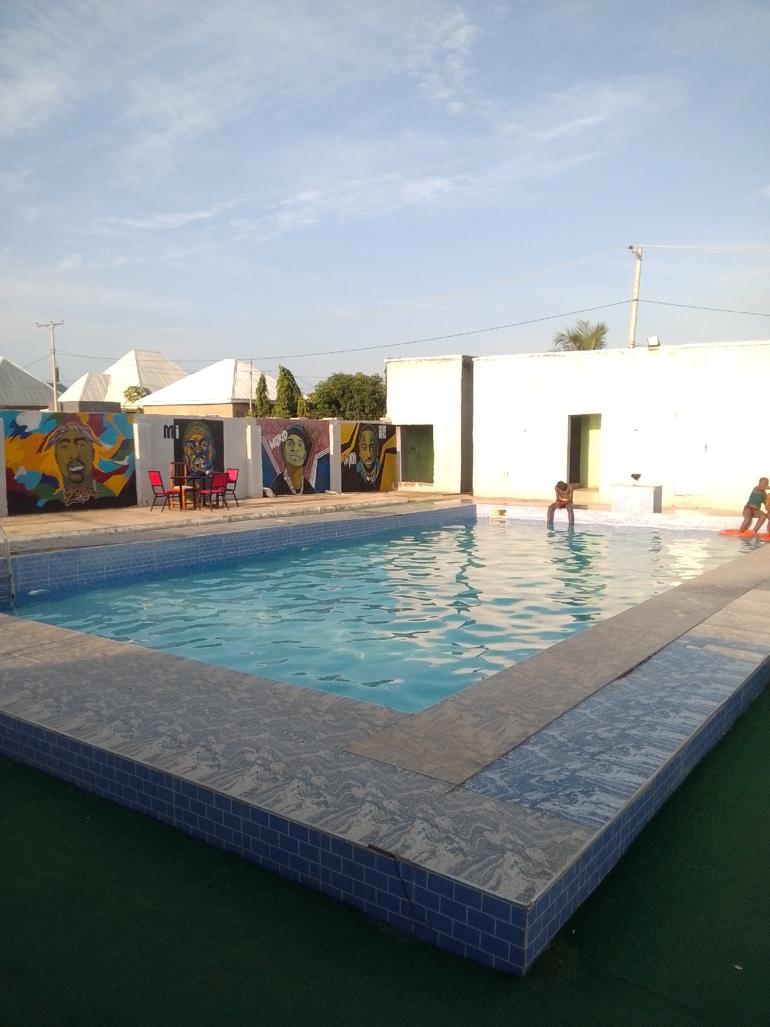 In Jalingo, Nigeria, a swimming pool area is depicted under a partly cloudy sky. The pool, filled with clear blue water, is surrounded by a tiled edge and a green artificial turf surface. The far wall of the pool area is adorned with large, colorful murals of various famous individuals. In front of the murals, a small table with four red chairs is set up.
Near the pool, on the right side of the image, two individuals are visible. One person, partially submerged in the water, is sitting on the edge of the pool, facing away from the camera. Further to the right, another person is floating on an orange inflatable device in the pool. A person is also visible near the left edge of the pool, appearing to climb out using the ladder.
The background shows white buildings with triangular roofs, a utility pole with wires, and trees. The overall scene suggests a recreational space, likely a hotel or a private residence, in Jalingo.